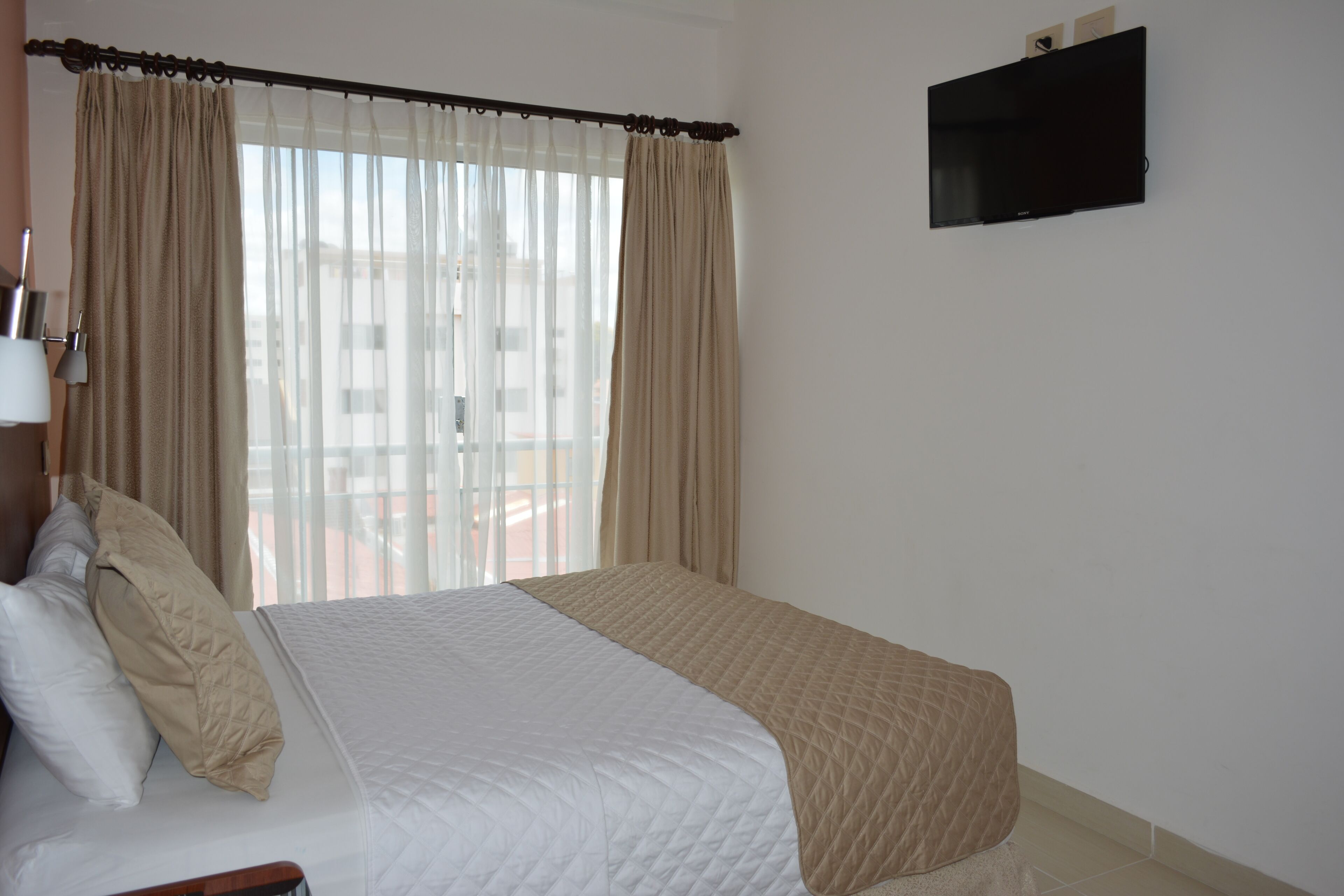 A hotel room with a bed, curtains, a TV on the wall, and a window with a view of buildings.