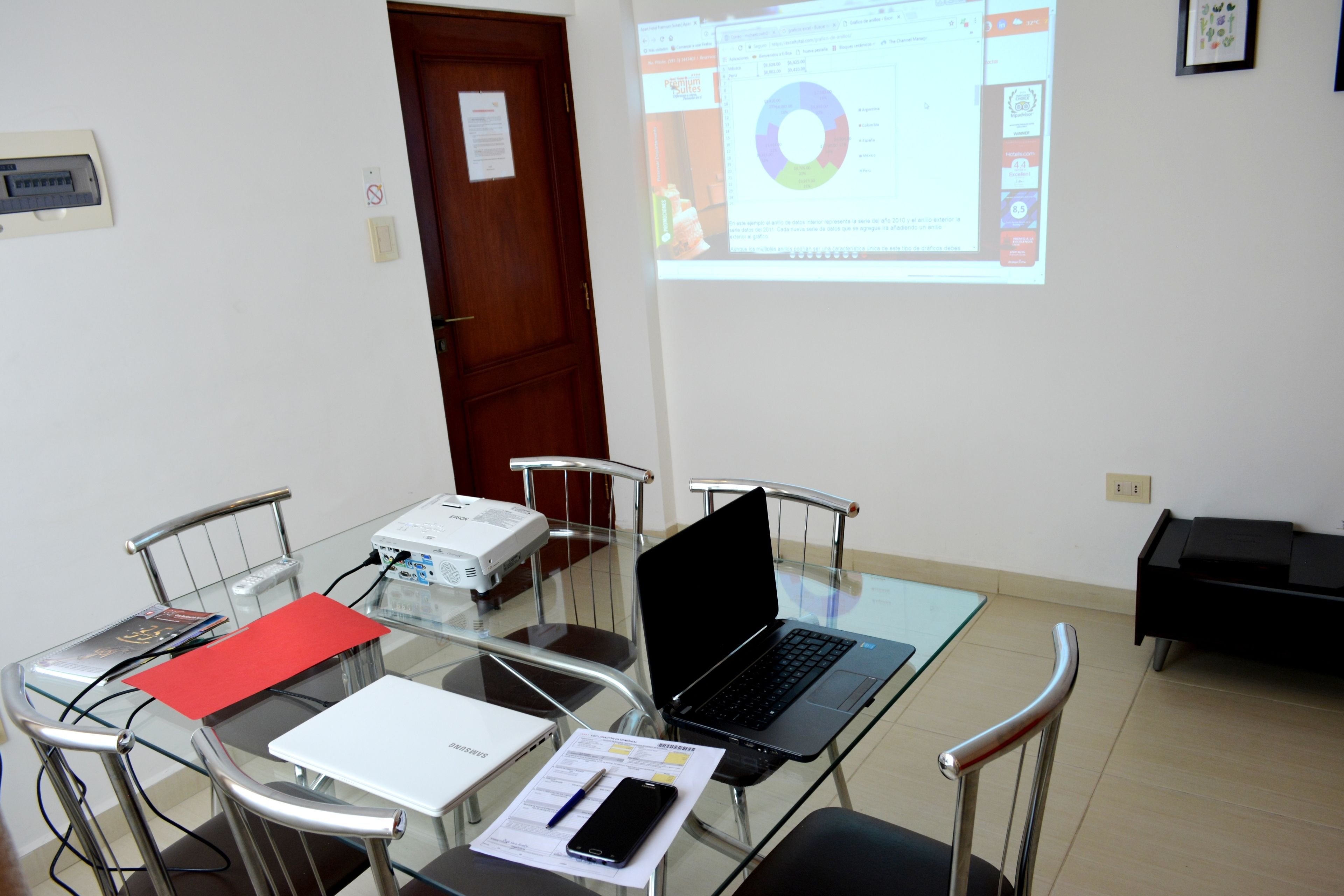 A conference room with a glass table, metal chairs, a laptop, a projector, and a presentation screen.