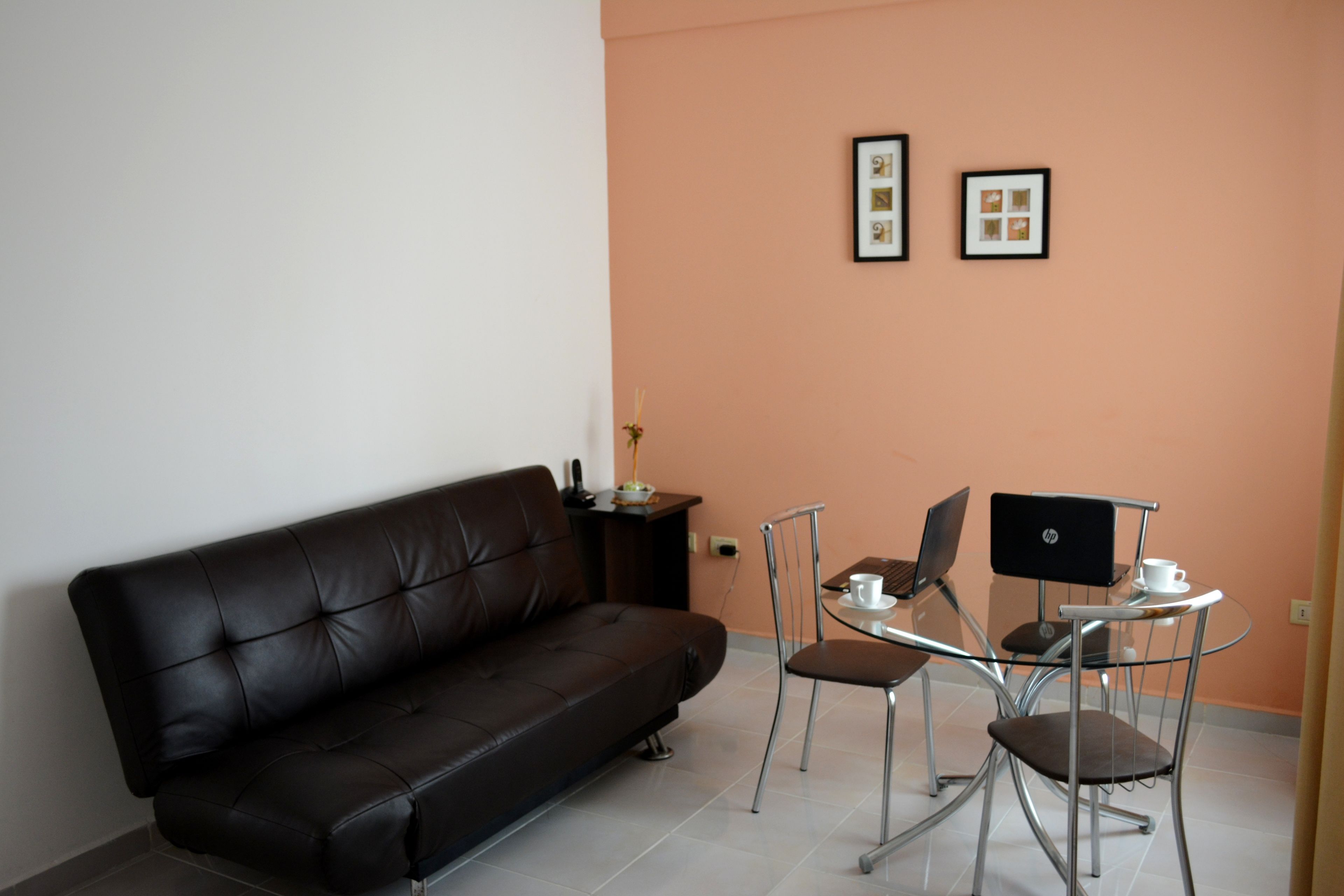 A modern living room with a black leather sofa, a glass table with two chairs, and a laptop.