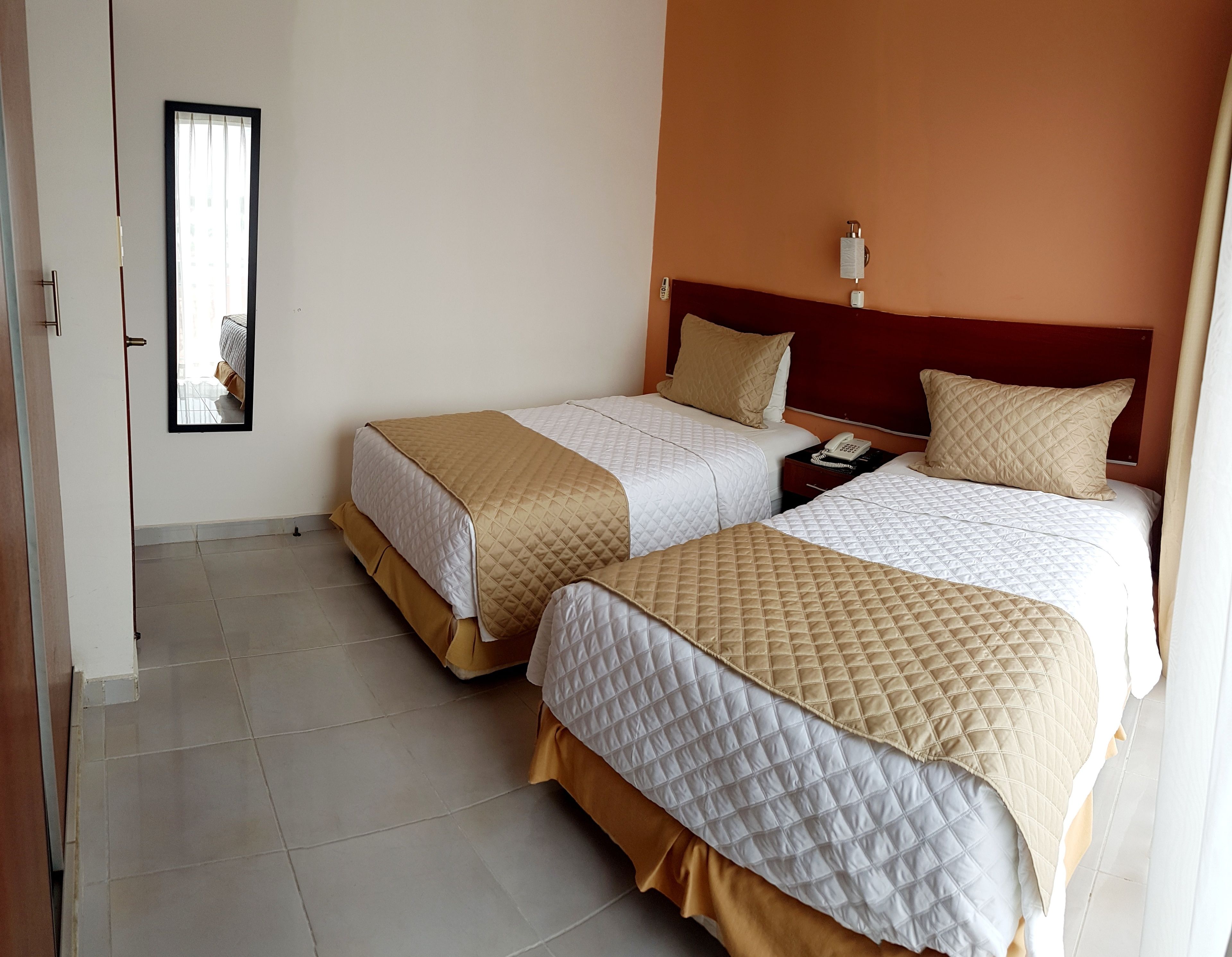 A hotel room with two beds, each with a quilted bedspread and pillows, a nightstand with a phone, and a wall-mounted light fixture.