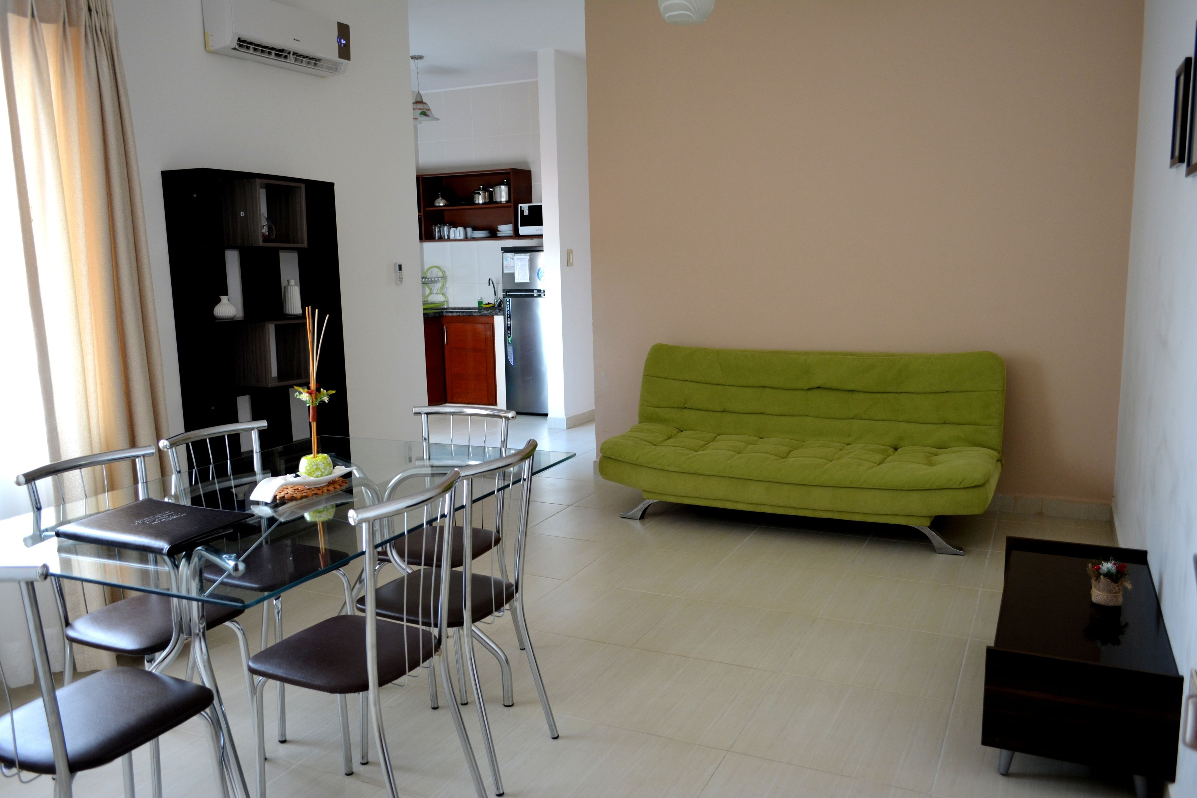 Een moderne woonkamer met een glazen eettafel, metalen stoelen, een groene bank en een keuken op de achtergrond.