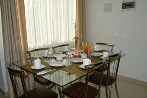 Un comedor con mesa de vidrio, sillas metálicas y una disposición para el desayuno, incluyendo platos, tazas y una jarra de jugo de naranja.