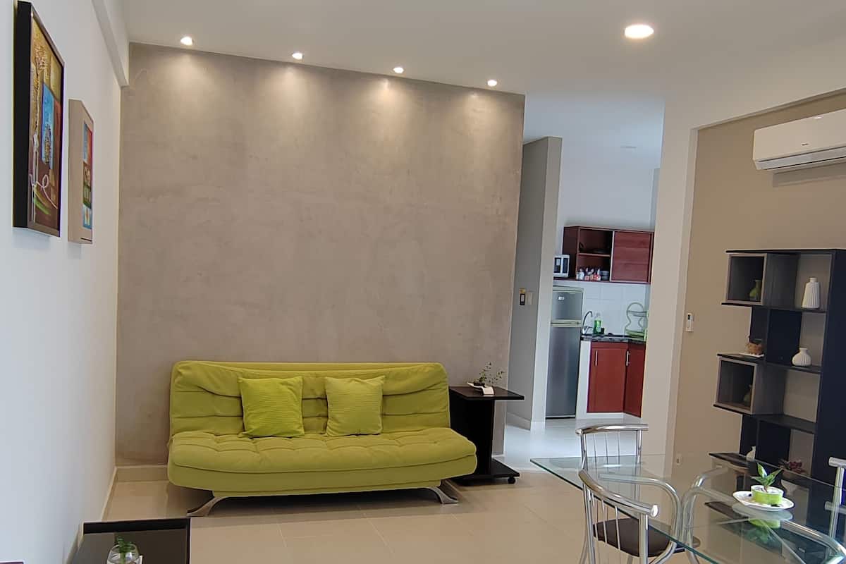 A modern living room with a green sofa, a glass dining table with chairs, a small side table, and a kitchen in the background.