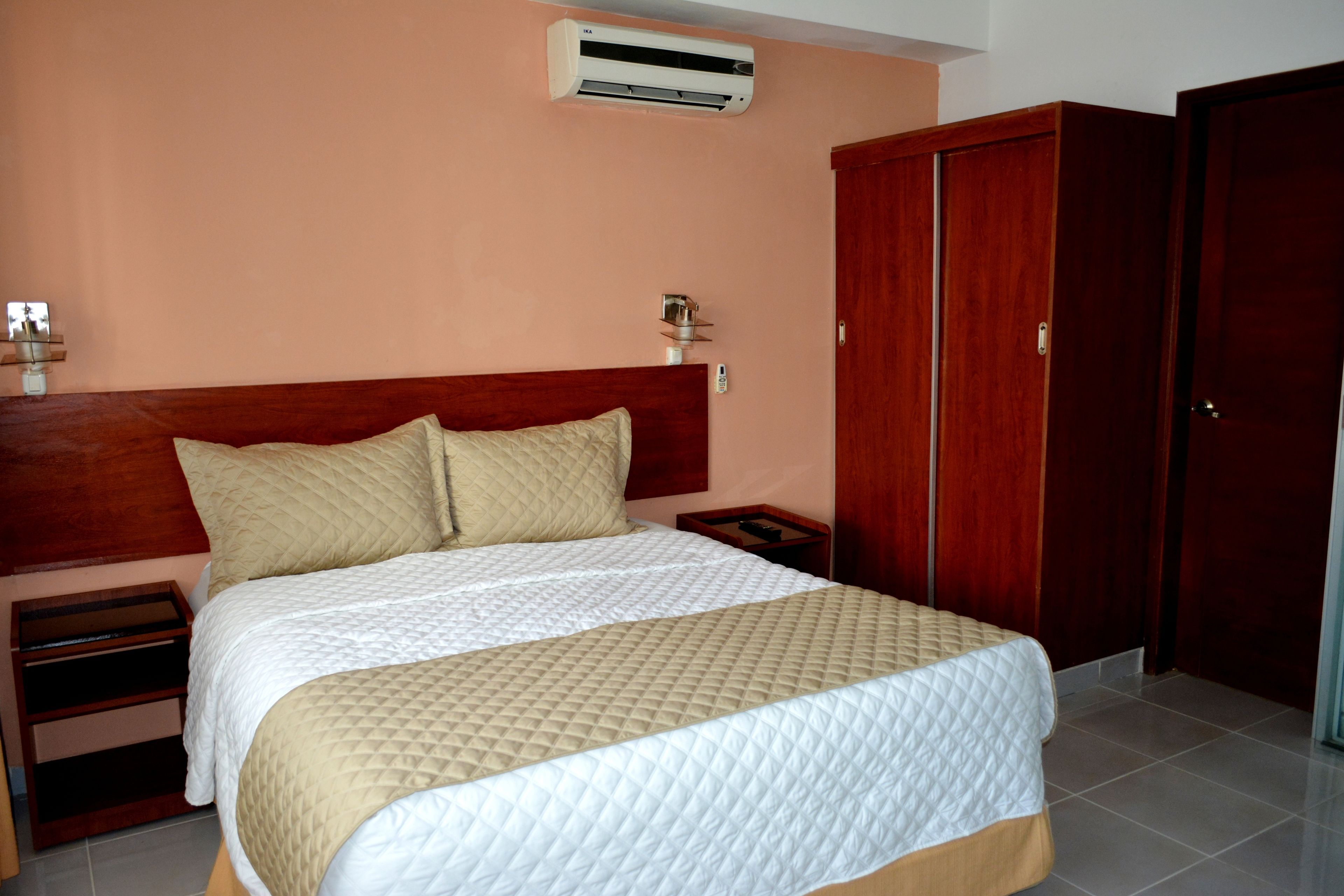 Una cama bien tendida con cubrecama acolchado, cabecera de madera, mesita de noche con lámpara y armario con espejo.