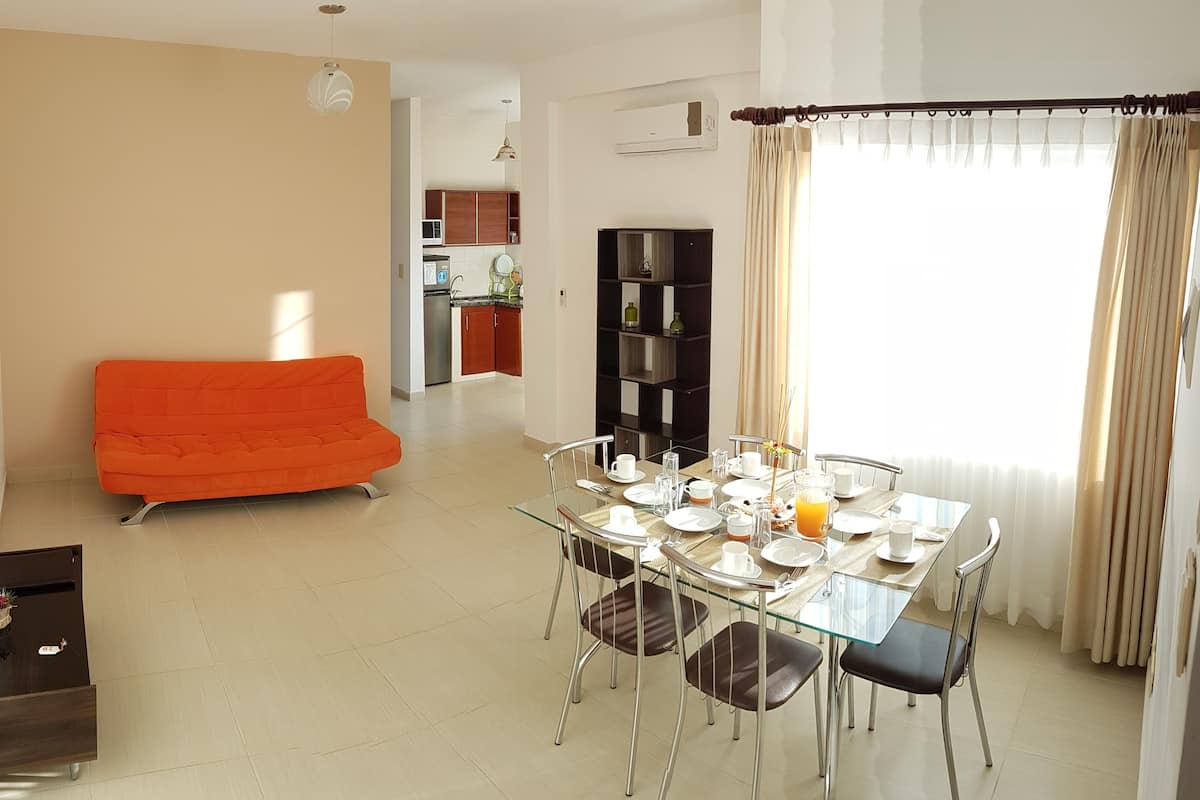 A dining area with a glass table, metal chairs, and a buffet setup. There is a red sofa in the living area and a kitchen in the background.