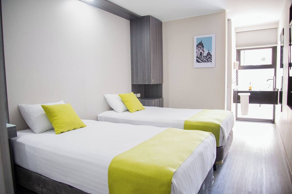 A hotel room with two beds, each with white bedding and a green throw. There is a wooden cabinet above the beds and a framed picture on the wall.