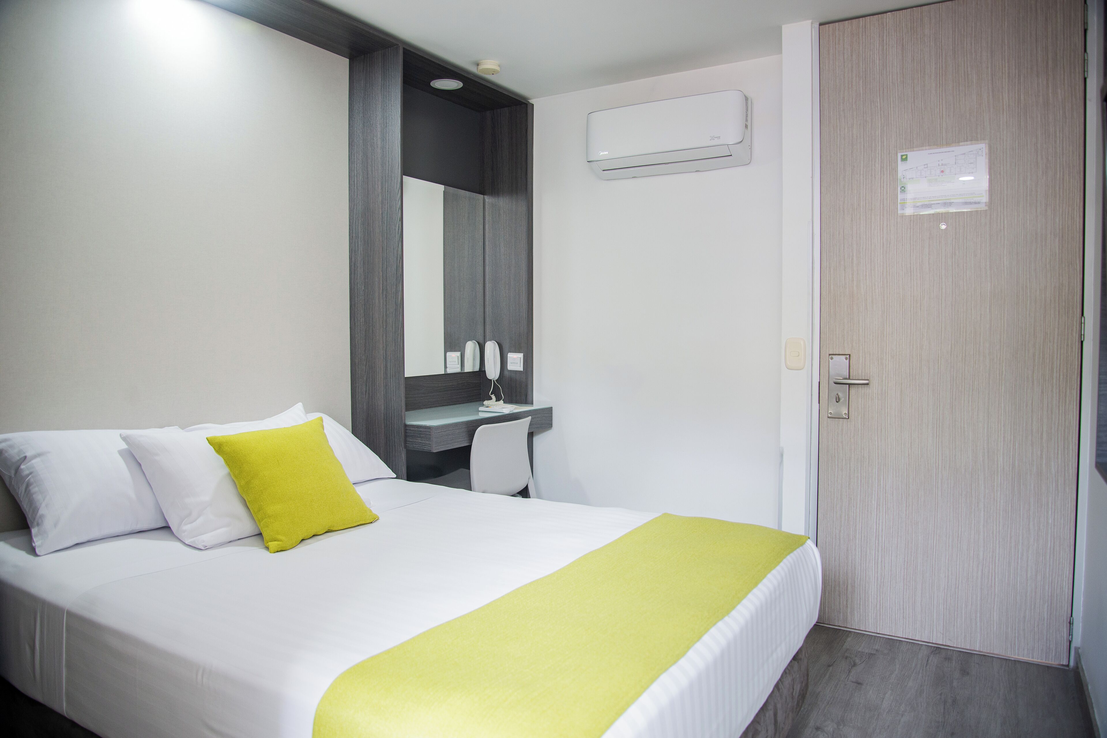 A hotel room with a bed, a desk, a chair, a mirror, an air conditioning unit, and a door.