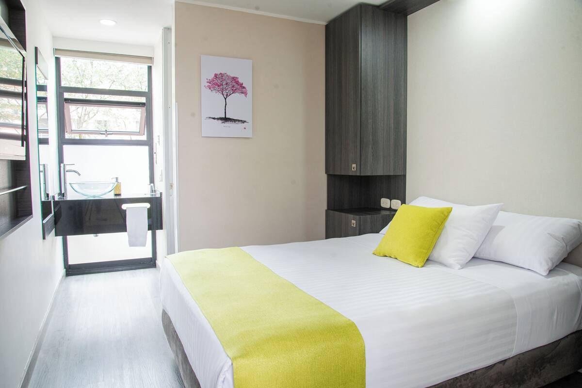 A modern hotel room with a bed, a bedside table, a mirror, and a window with a view.