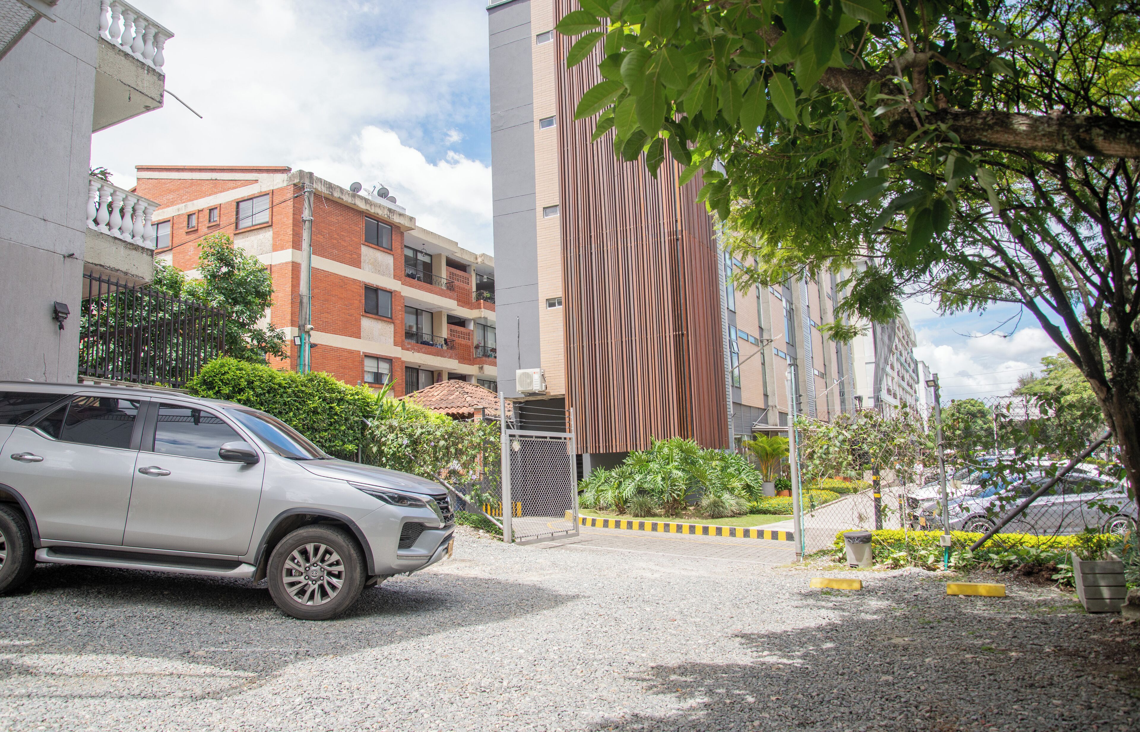 A parking area with a silver SUV, a building with a modern facade, and a tree in the foreground.