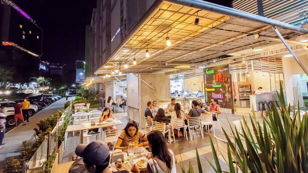 An outdoor dining area with tables and chairs, a pizza restaurant in the background, and a street with cars and people.