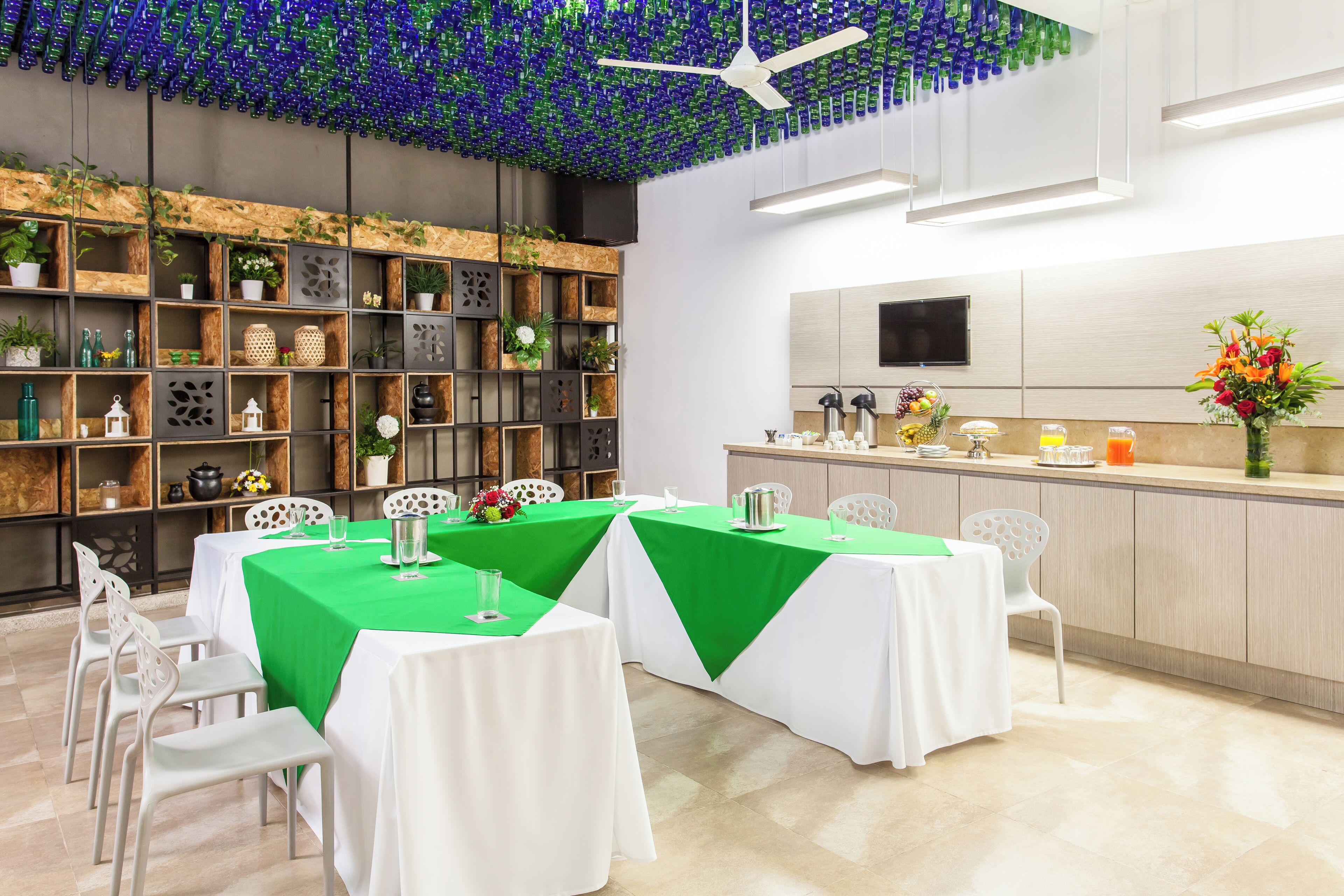 Un espace repas avec des tables disposées pour accueillir quatre personnes, un coin bar équipé d’un téléviseur et un plafond décoré de bouteilles bleues et vertes.