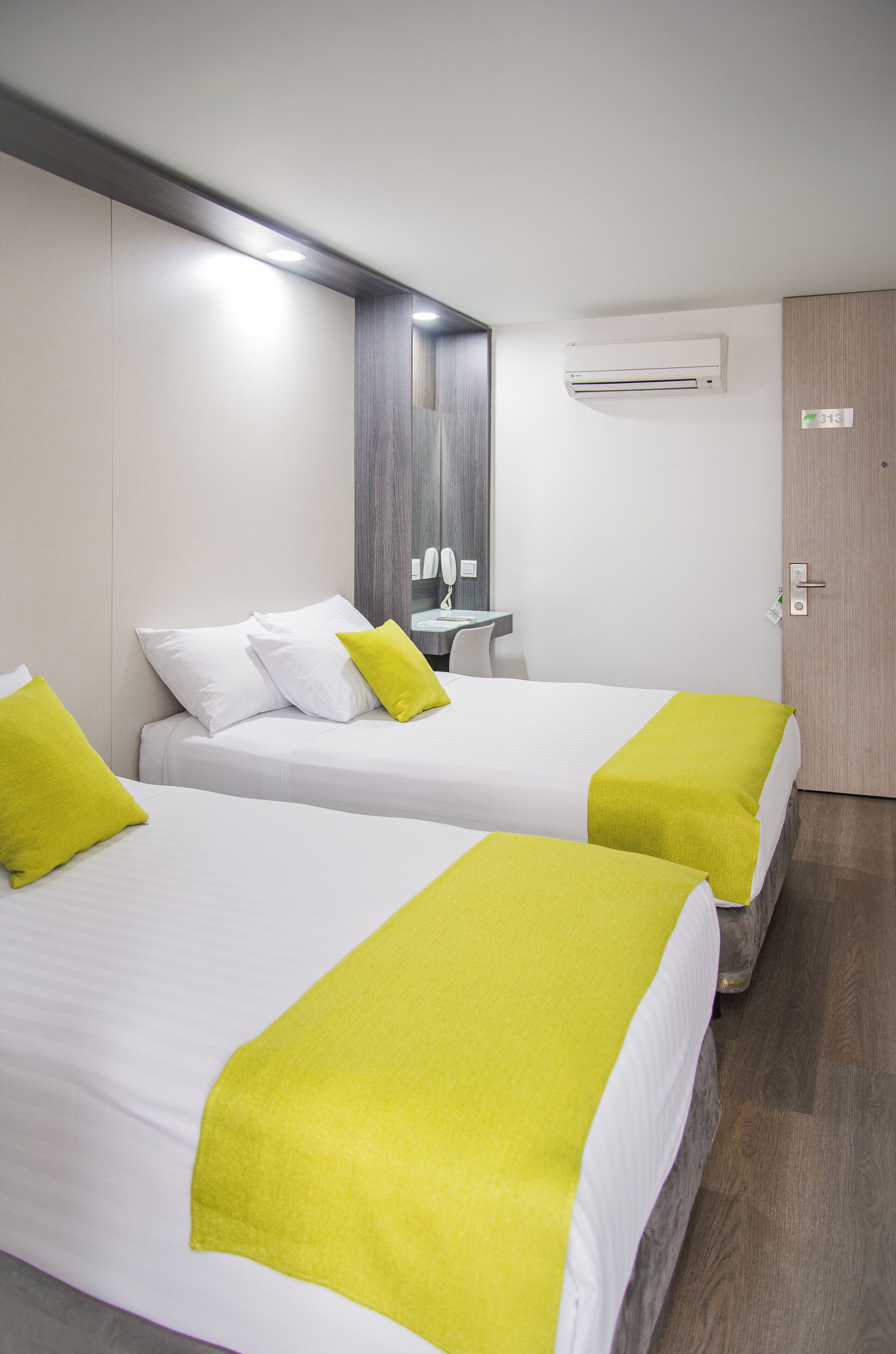 A hotel room with two beds, each with white bedding and a bright yellow accent pillow. There is a small bedside table with a lamp, a mirror, and a wall-mounted air conditioning unit.
