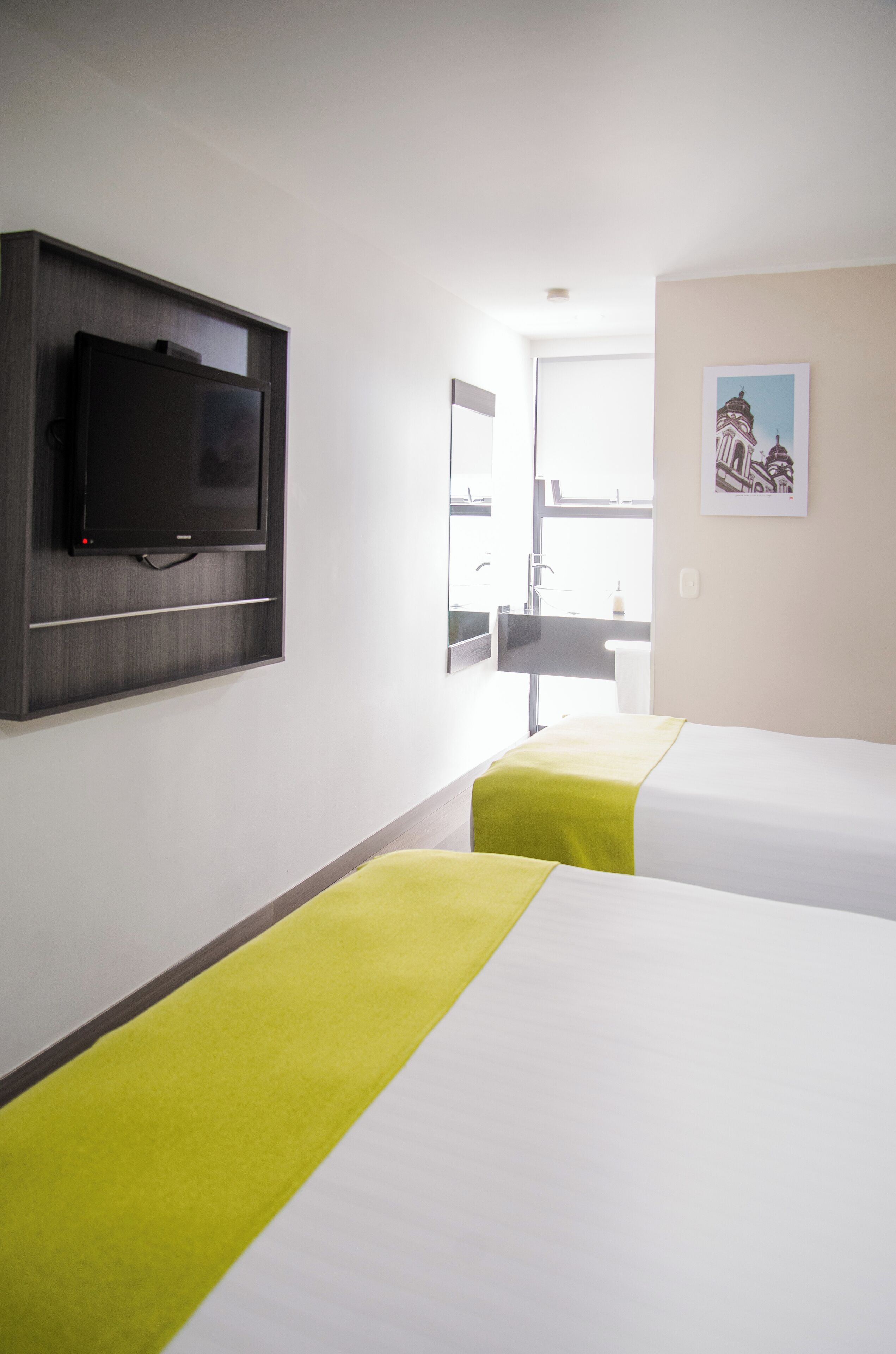A hotel room with two beds, a flat-screen TV mounted on the wall, a window with a view, and a framed picture on the wall.