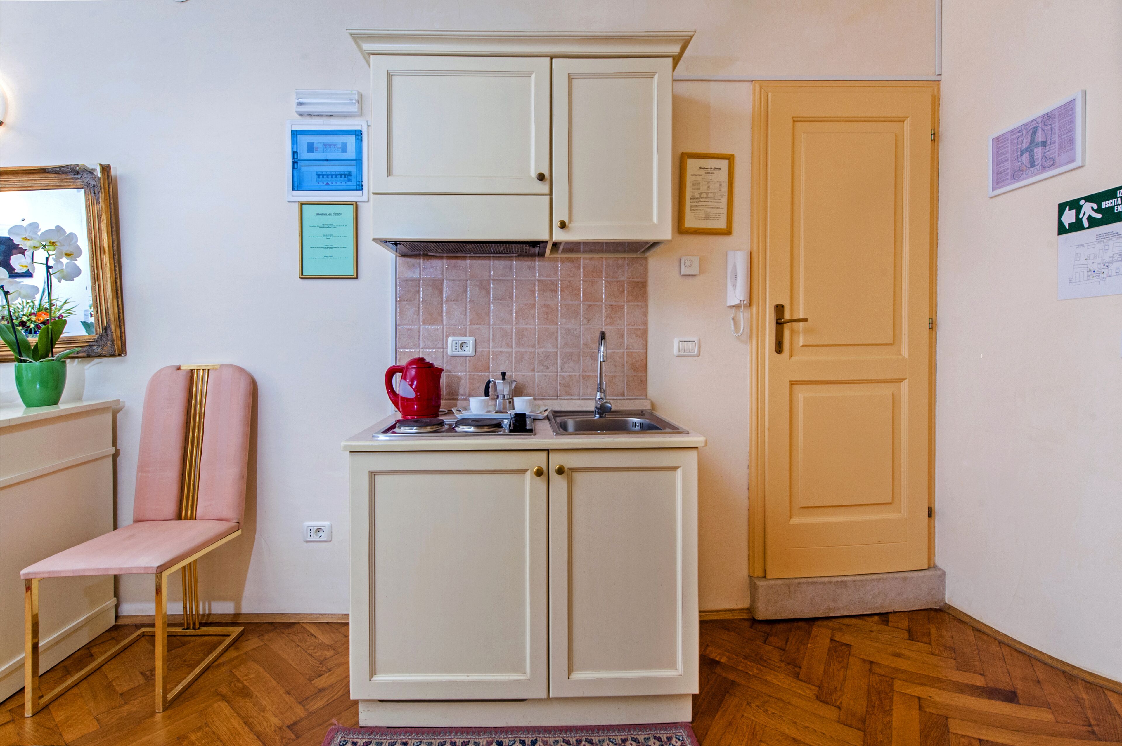 A compact kitchen with white cabinets, a sink, and a small countertop. There is a chair and a framed picture on the wall.