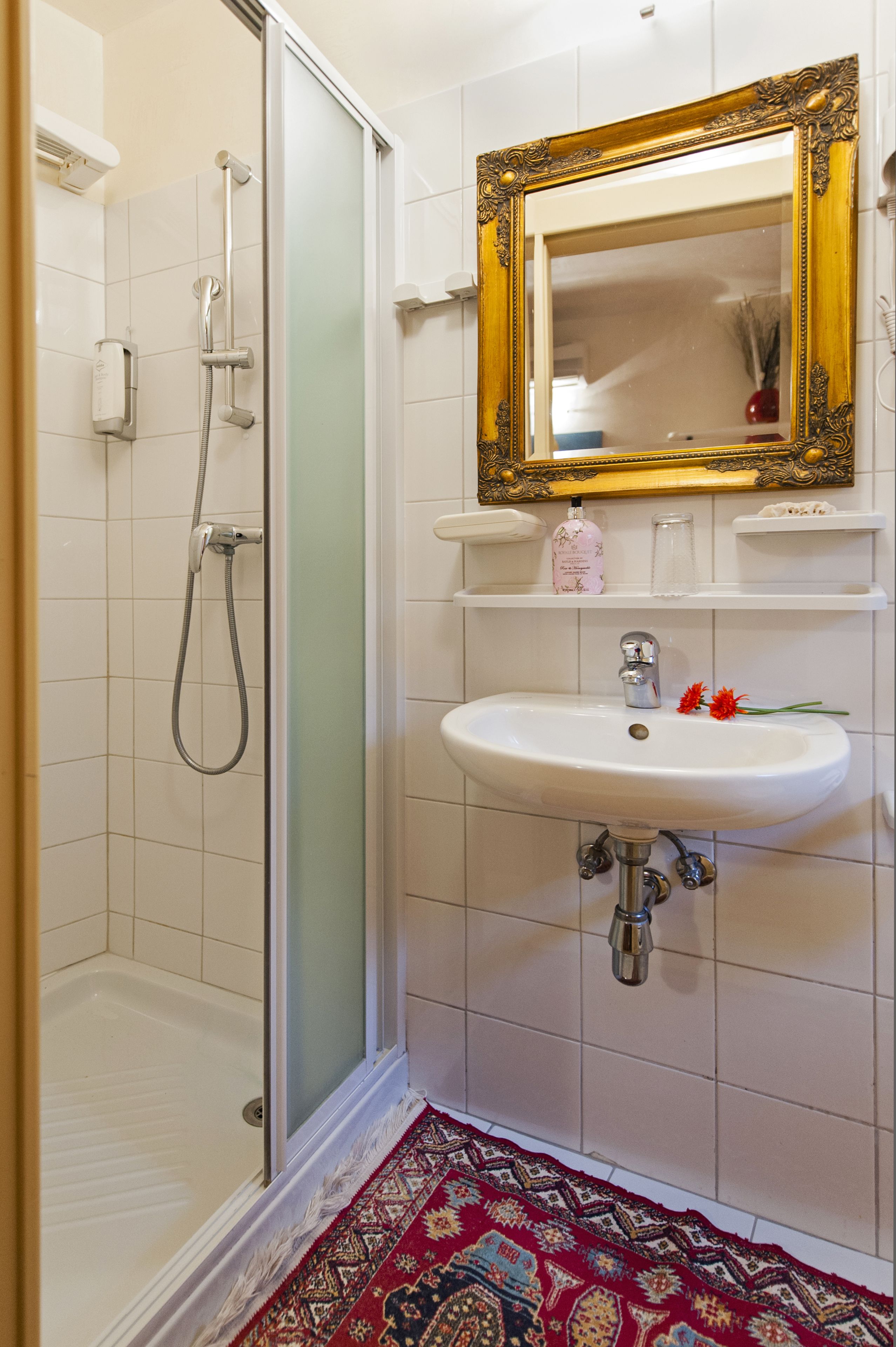 A bathroom with a glass shower enclosure, a white sink with a gold-framed mirror above it, and a patterned rug on the floor.