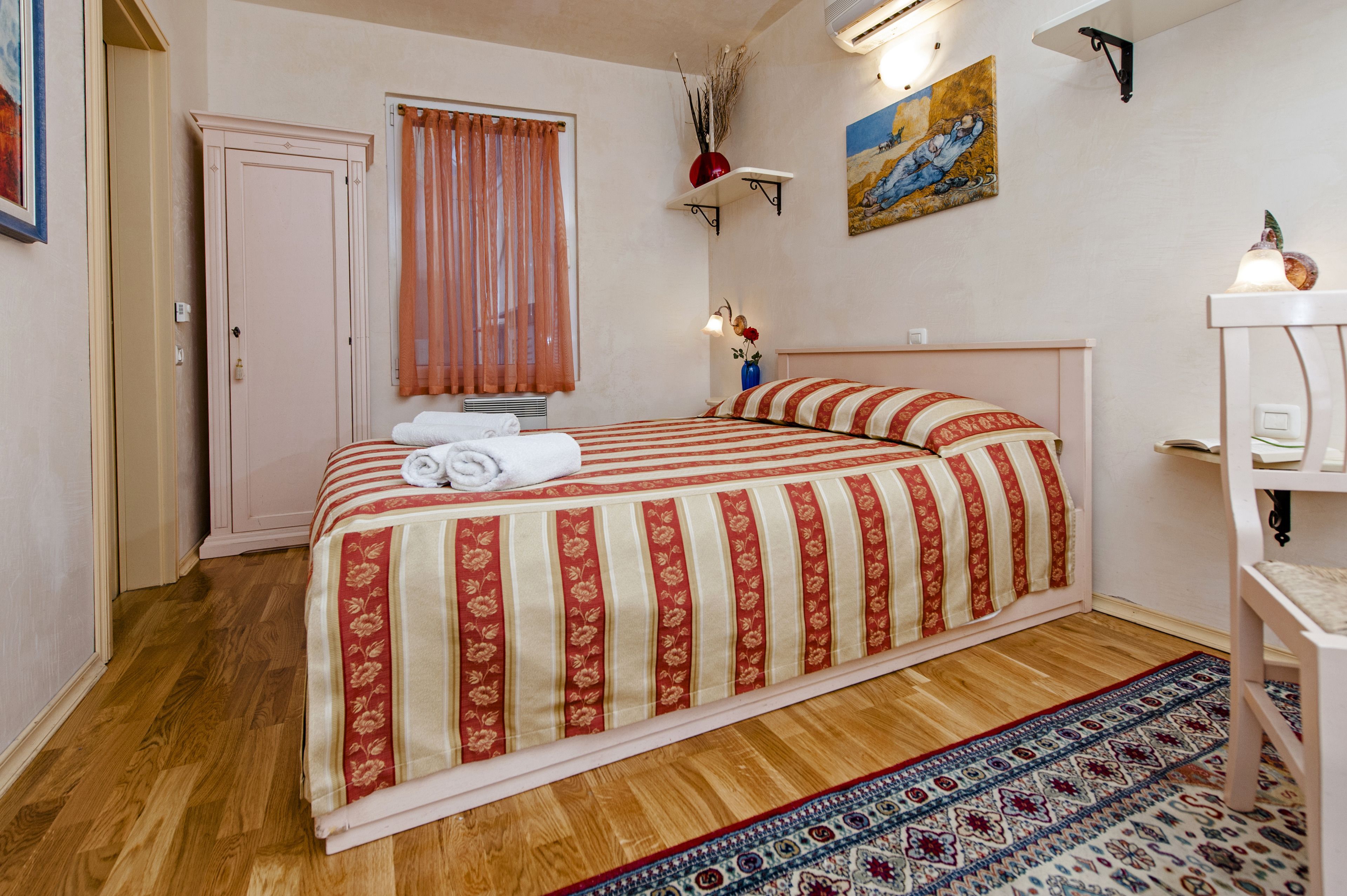 A bedroom with a bed, a nightstand, a window with curtains, a painting on the wall, and a wooden floor.