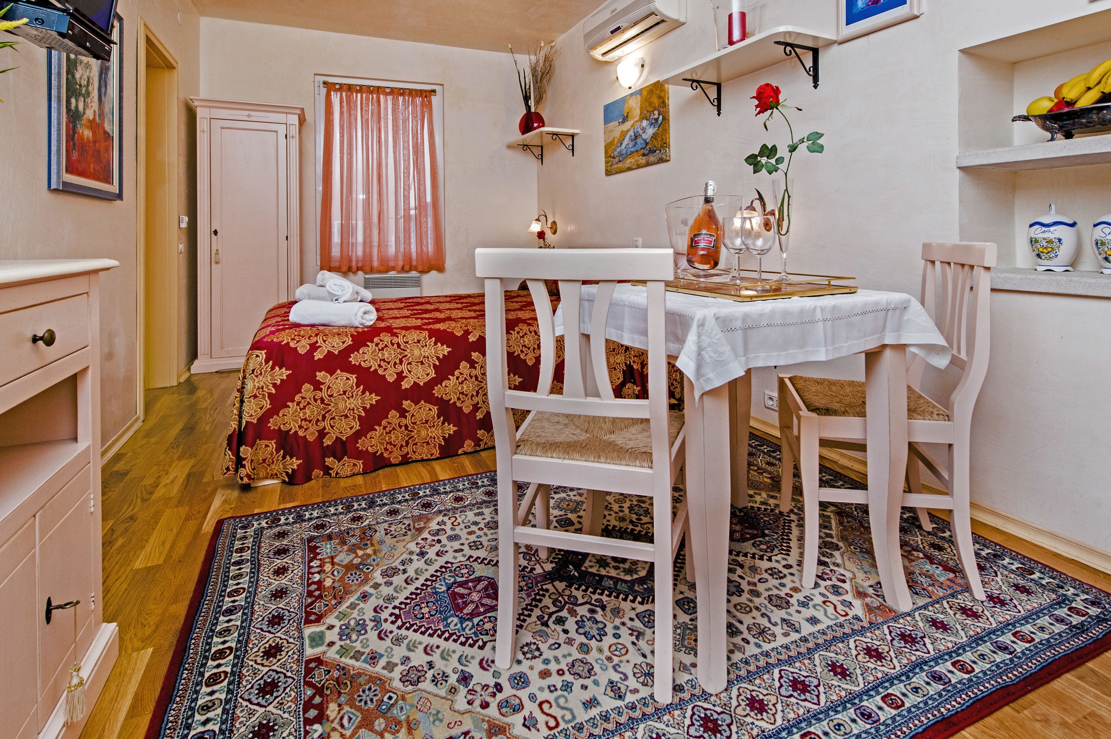 A hotel room with a bed, a dining table with chairs, a small table with a bottle and glasses, a shelf with decorative items, and a patterned rug.