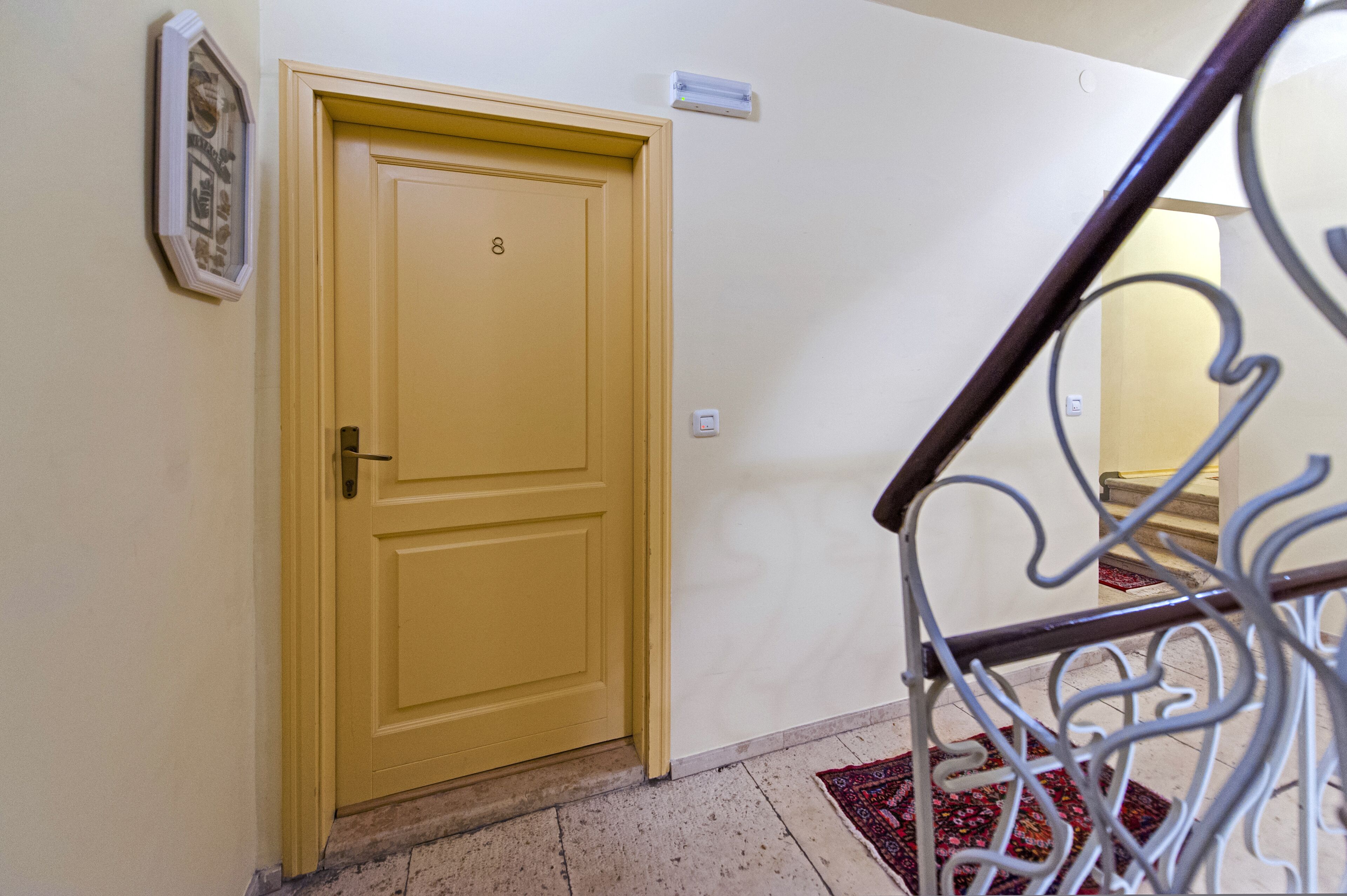 A yellow door with the number 8, a white wall, a white light switch, and a red patterned rug.