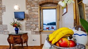 A room with a stone wall, a television mounted on it, a window with a view of boats, a decorative vase with flowers, and a fruit bowl on the table. - Residence La Carera (Rovinj)