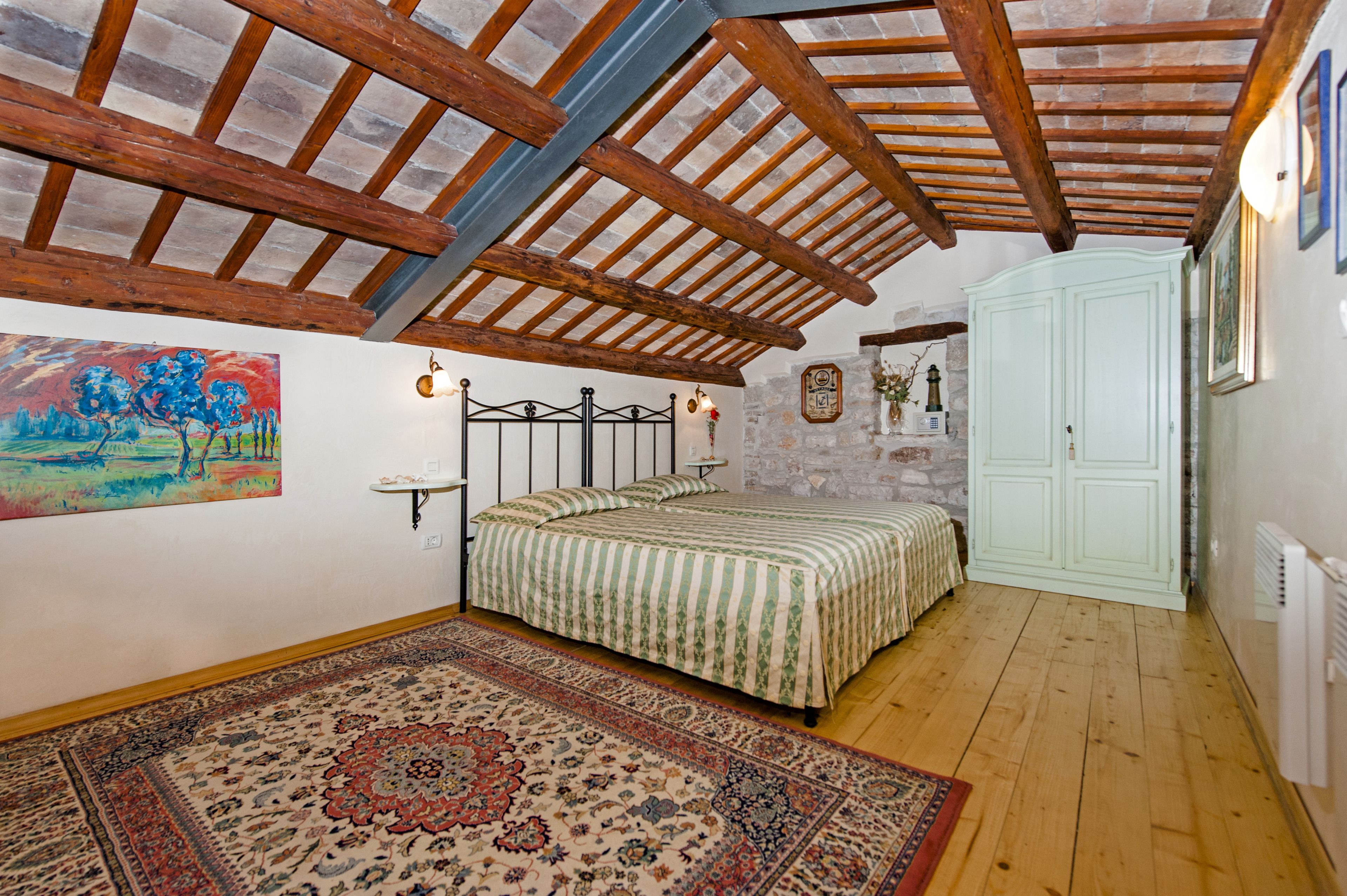 Une chambre avec un plafond en bois, un lit recouvert d’une couvre-lit à motifs, un mur en pierre et un grand tapis.