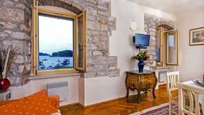 A room with a stone wall, a window with an open shutter, a television, a small table with a vase, a white chair, and a patterned rug. - Residence La Carera (Rovinj)