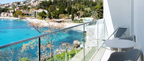 A balcony with a glass railing overlooking a coastal area with clear blue waters and a view of houses on a hillside.