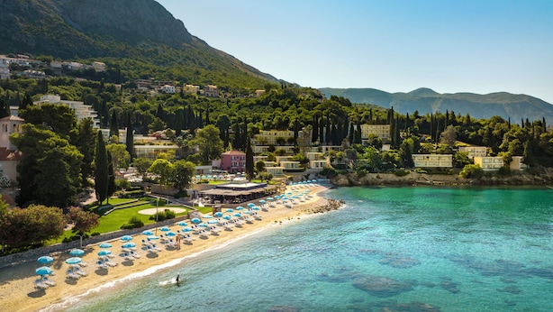 A coastal resort with a sandy beach, clear turquoise waters, and a backdrop of lush green hills.