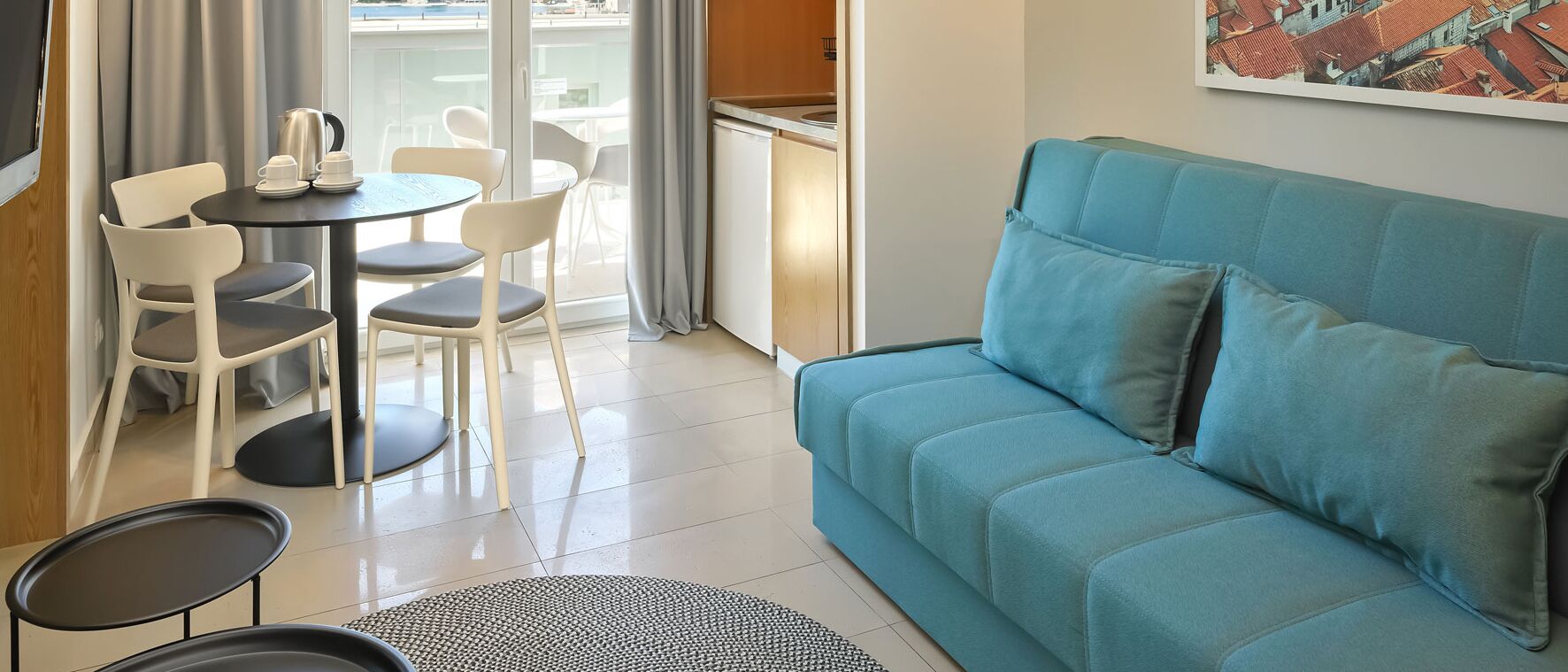 Ein modernes Wohnzimmer mit einem türkisblauen Sofa, einem kleinen runden Tisch mit Stühlen und Blick auf einen Balkon mit Stadtansicht.