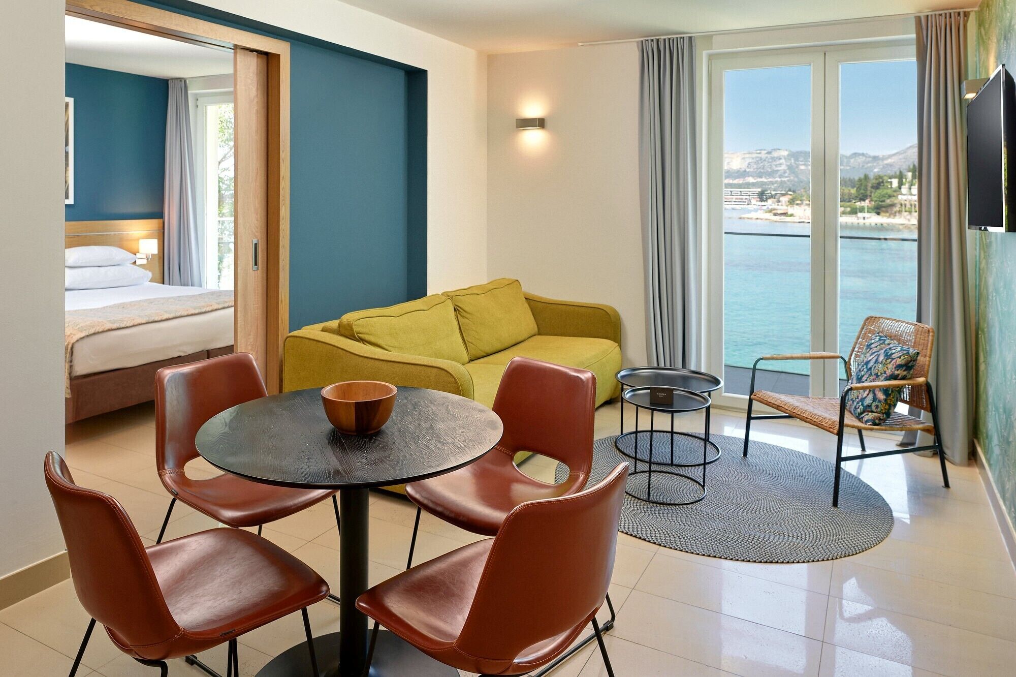 Habitación de hotel con cama, sofá amarillo, mesa de comedor con sillas, televisor y vista al mar.