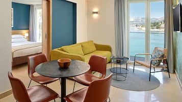A hotel room with a bed, a yellow sofa, a dining table with chairs, a TV, and a view of the sea.