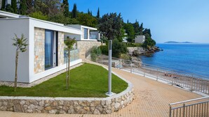 A modern coastal property with a stone facade, a grassy lawn, and a paved walkway leading to the sea.