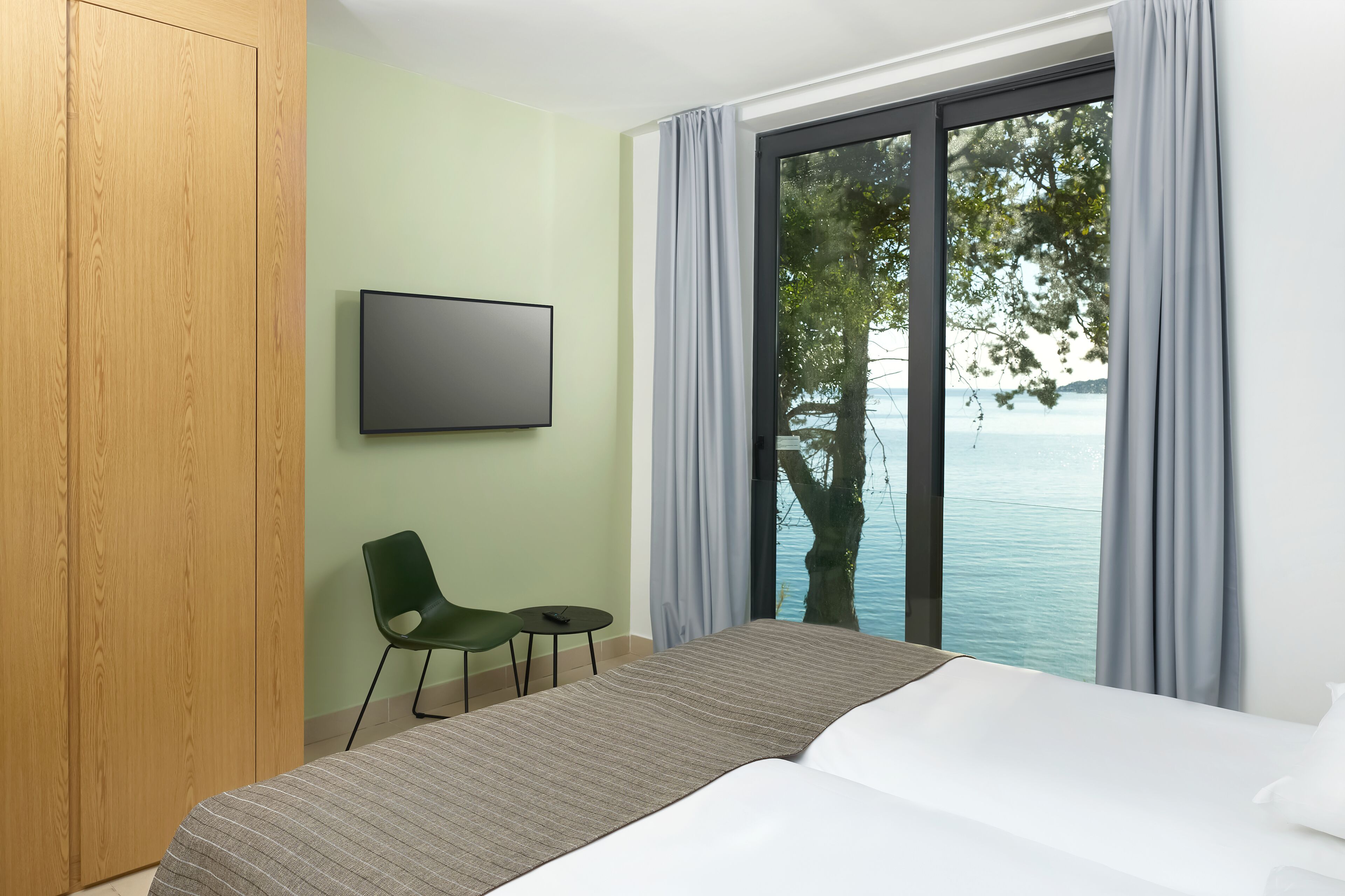 A hotel room with a bed, a television, a chair, and a view of the sea.