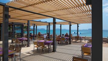 An outdoor dining area with tables and chairs, wooden pergolas, and a view of the sea.