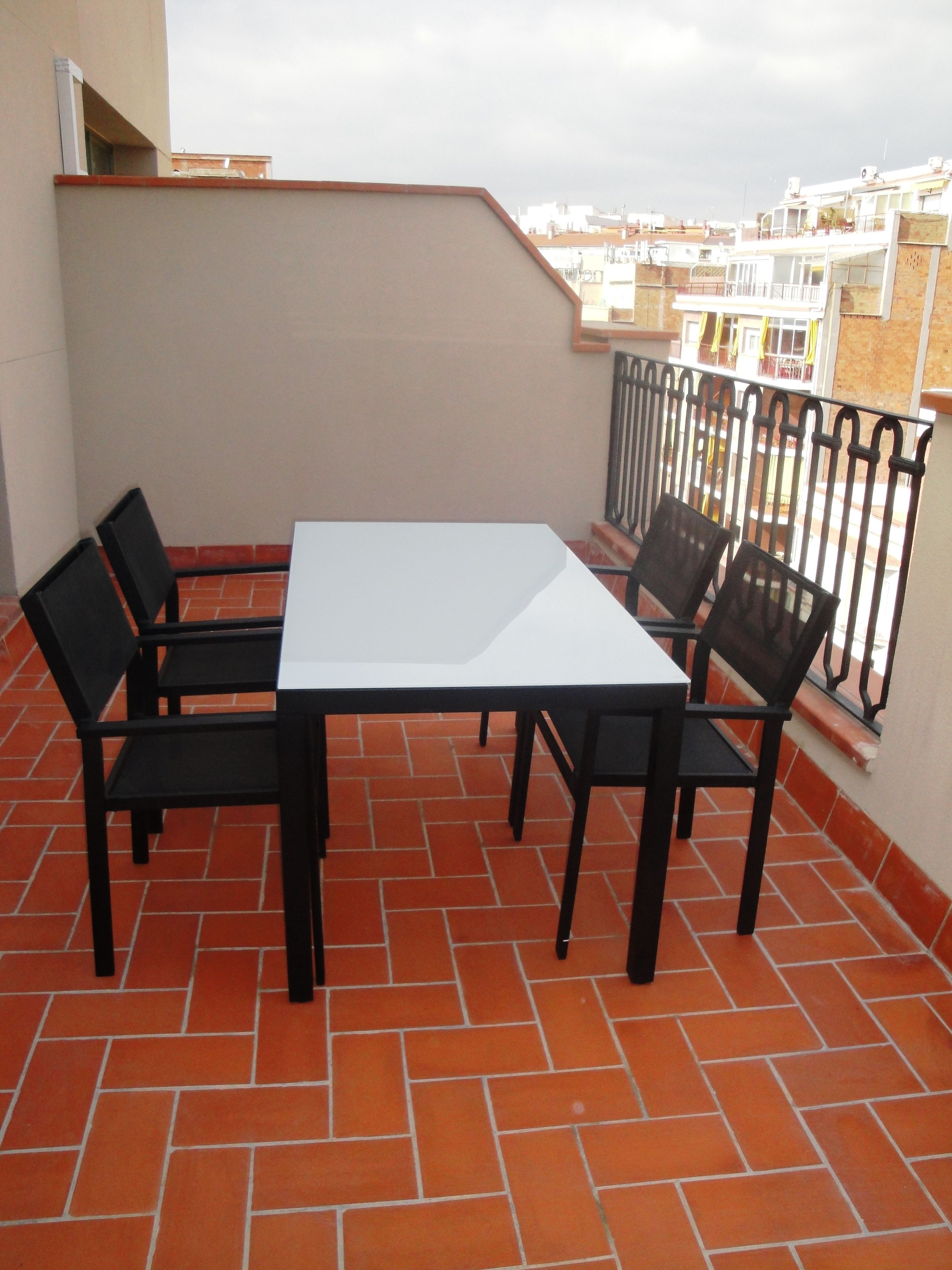 A terrace with a white table and four chairs, surrounded by a red-tiled floor and a metal railing.