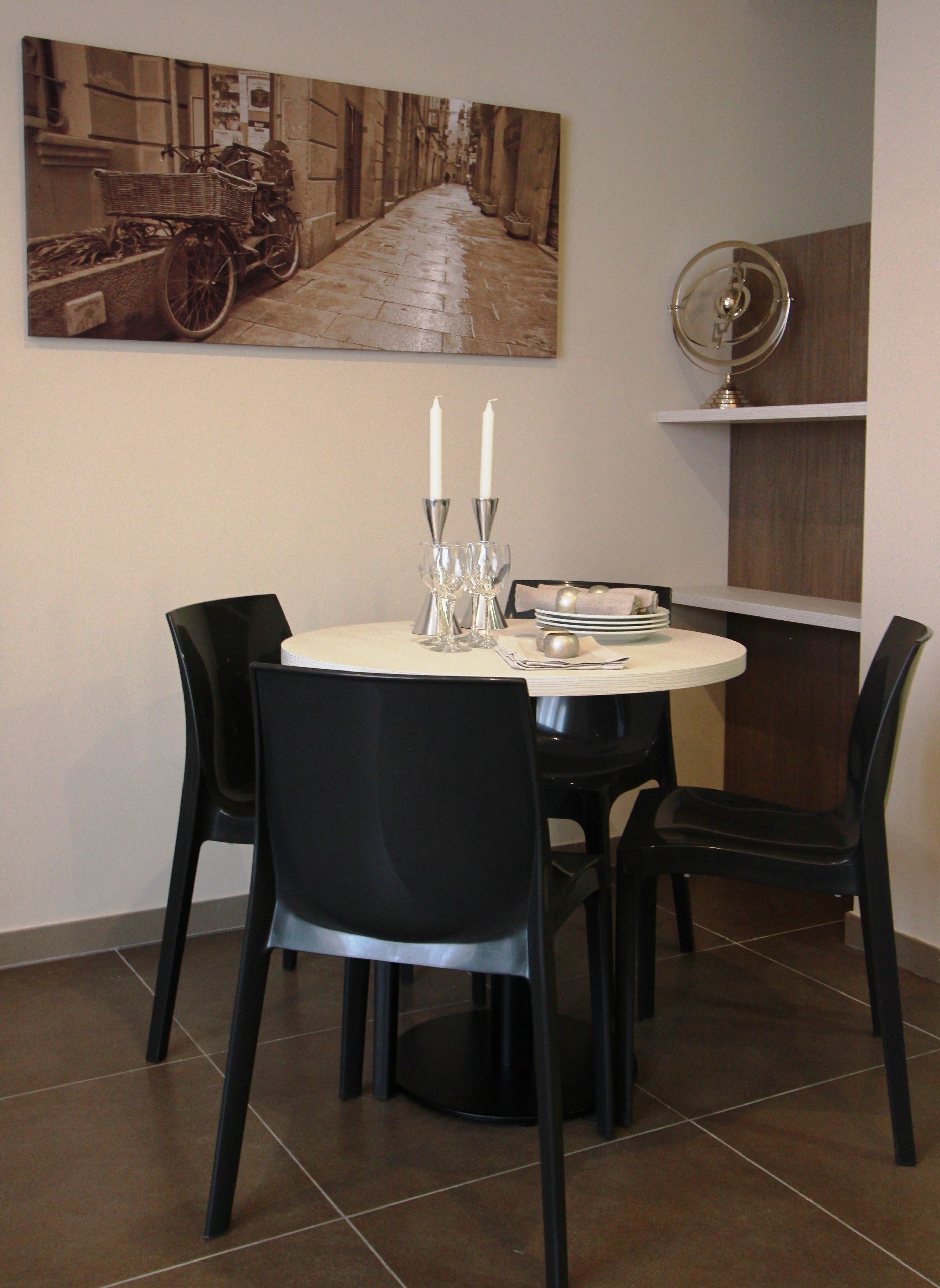 Un coin repas avec une table ronde, des chaises noires, un tableau au-dessus de la table et une petite étagère sur laquelle se trouve un ventilateur.