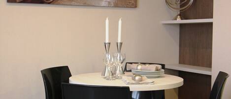 Un coin repas avec une table ronde, des chaises noires, un tableau au-dessus de la table et une petite étagère sur laquelle se trouve un ventilateur.