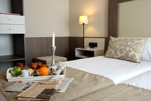 Ein Hotelzimmer mit einem ordentlich bezogenen Bett, einem Nachttisch mit Lampe, einem Tablett mit Frühstücksgebäck und einer Zeitung auf dem Bett.