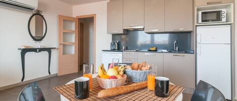 A modern kitchen with a dining table set for breakfast, including fruits, bread, and beverages.