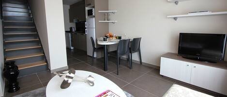 A modern living room with a dining area, a kitchen in the background, and a staircase leading to another level.