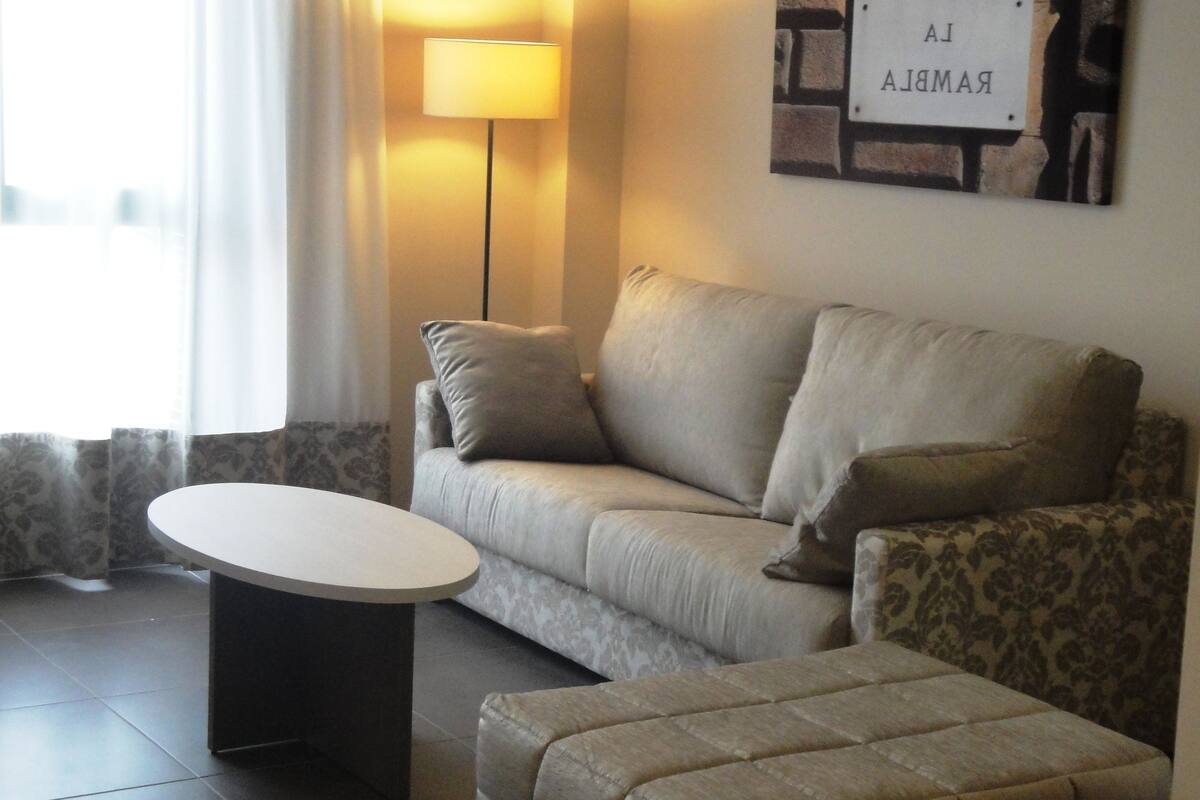 Un salon comprenant un canapé, un pouf et une table basse ronde. On y trouve également une lampe, un panneau décoratif fixé au mur et une fenêtre avec des rideaux.