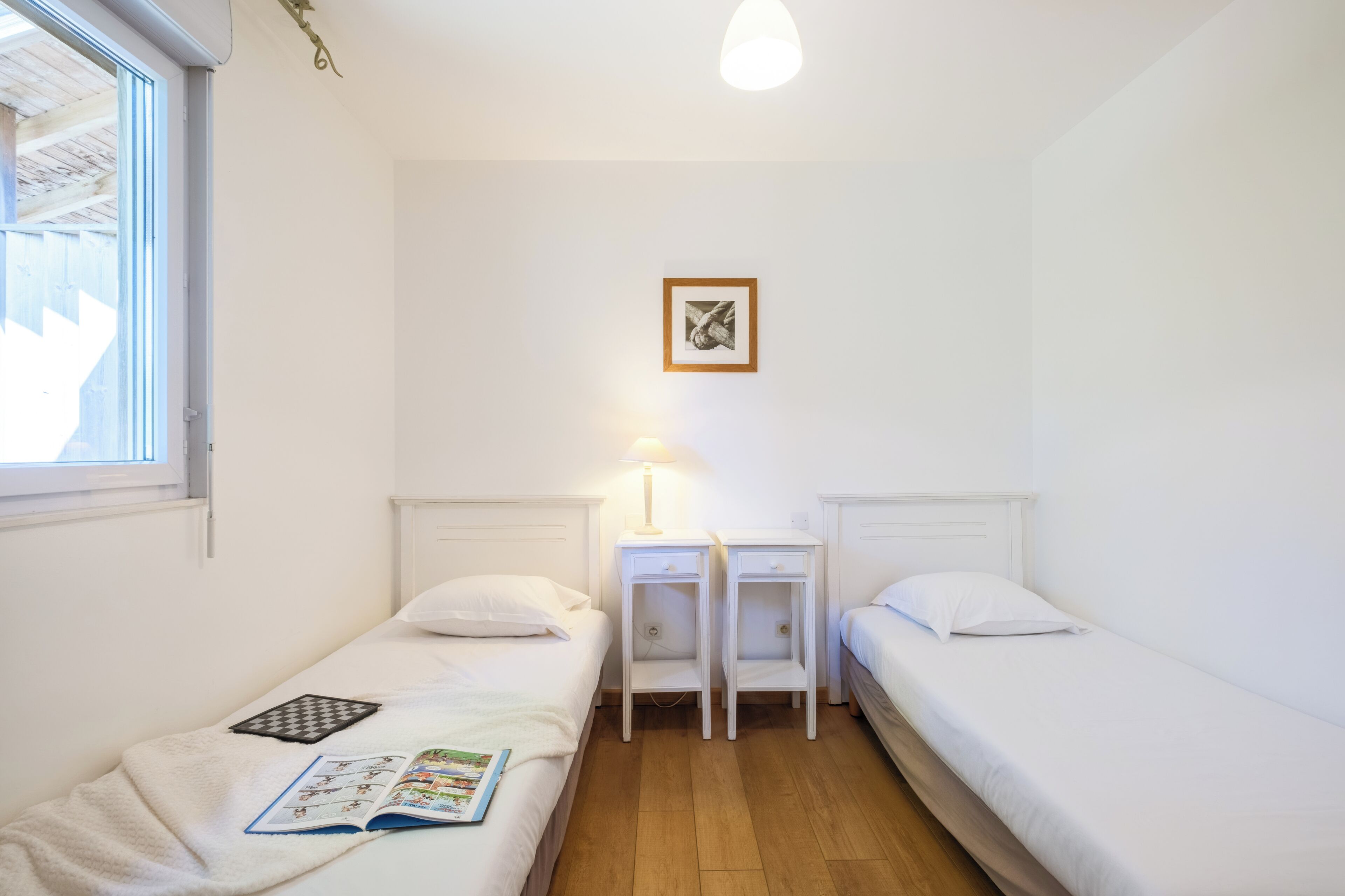 A small, clean room with two single beds, a bedside table with a lamp, a framed picture on the wall, and a window.