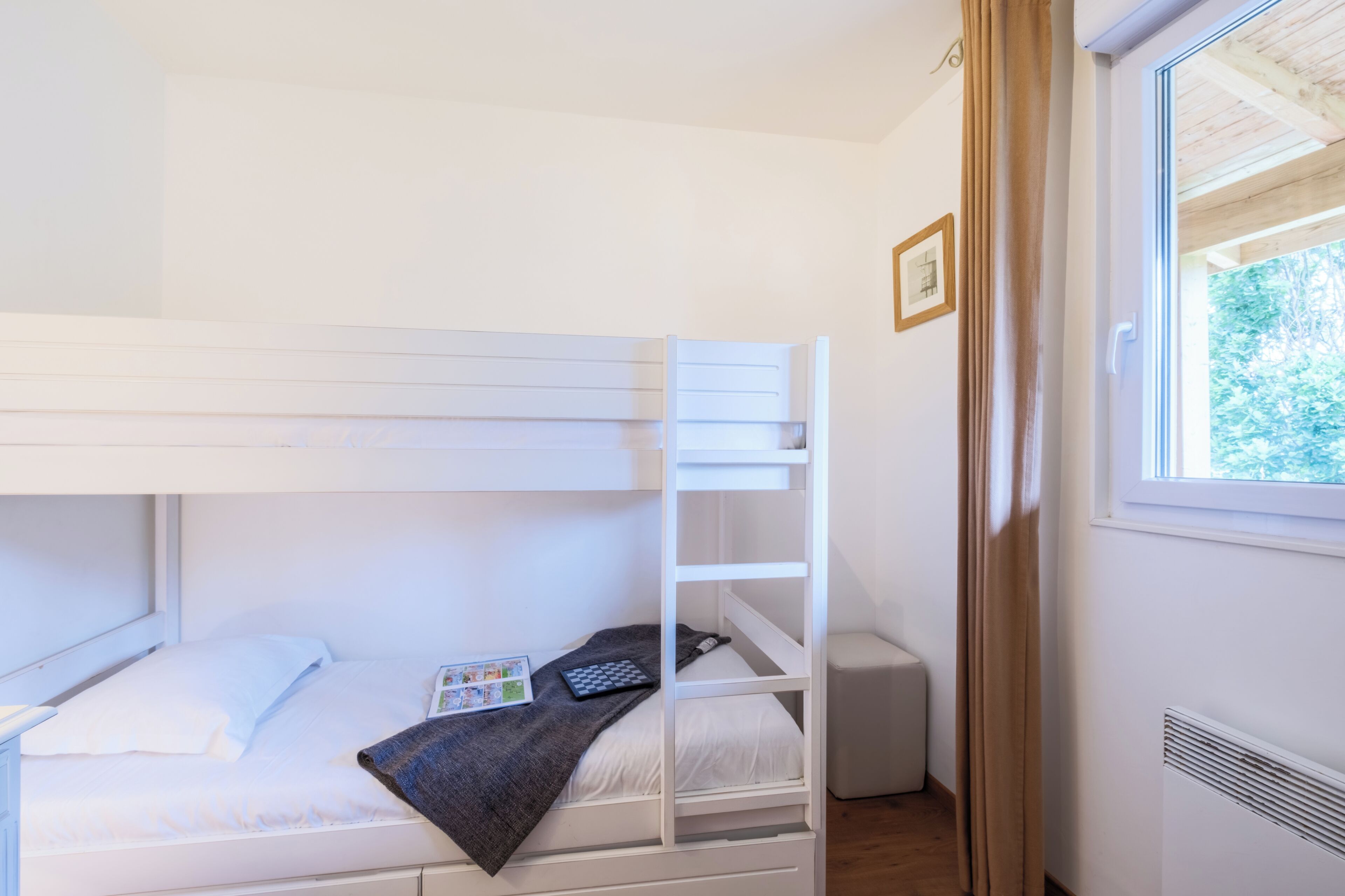 Una cama litera con una escalera, una mesita de noche con un libro y un mantel a cuadros, y una ventana con vista a follaje.