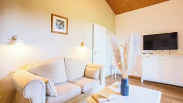 Un salon comprenant un canapé, une table basse avec un livre et un vase rempli d’herbe des pampas, un téléviseur et un meuble blanc.