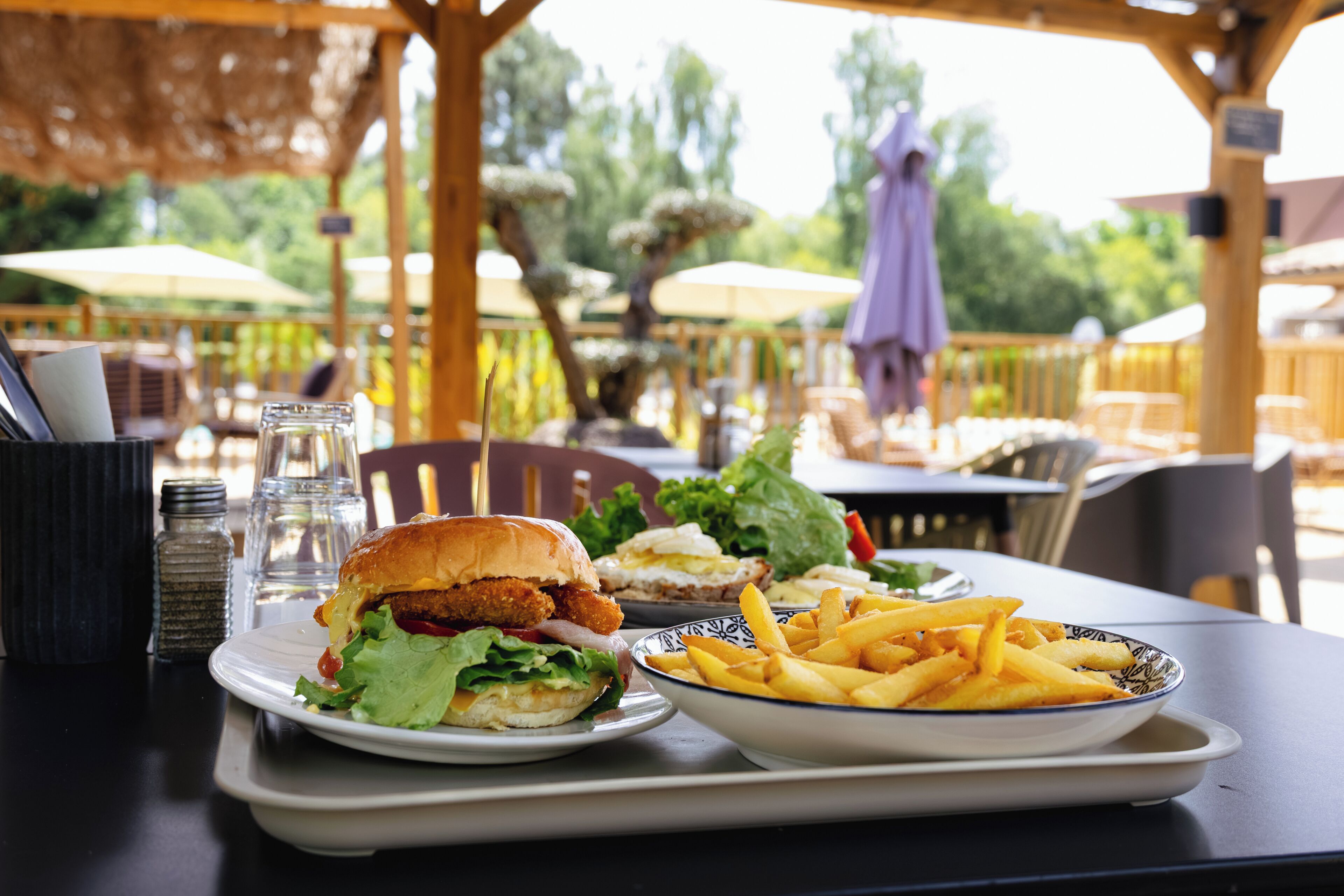 Eine Mahlzeit mit einem Burger, Pommes und einem Salat auf einem Tablett, im Hintergrund Sitzgelegenheiten im Freien und Sonnenschirme.