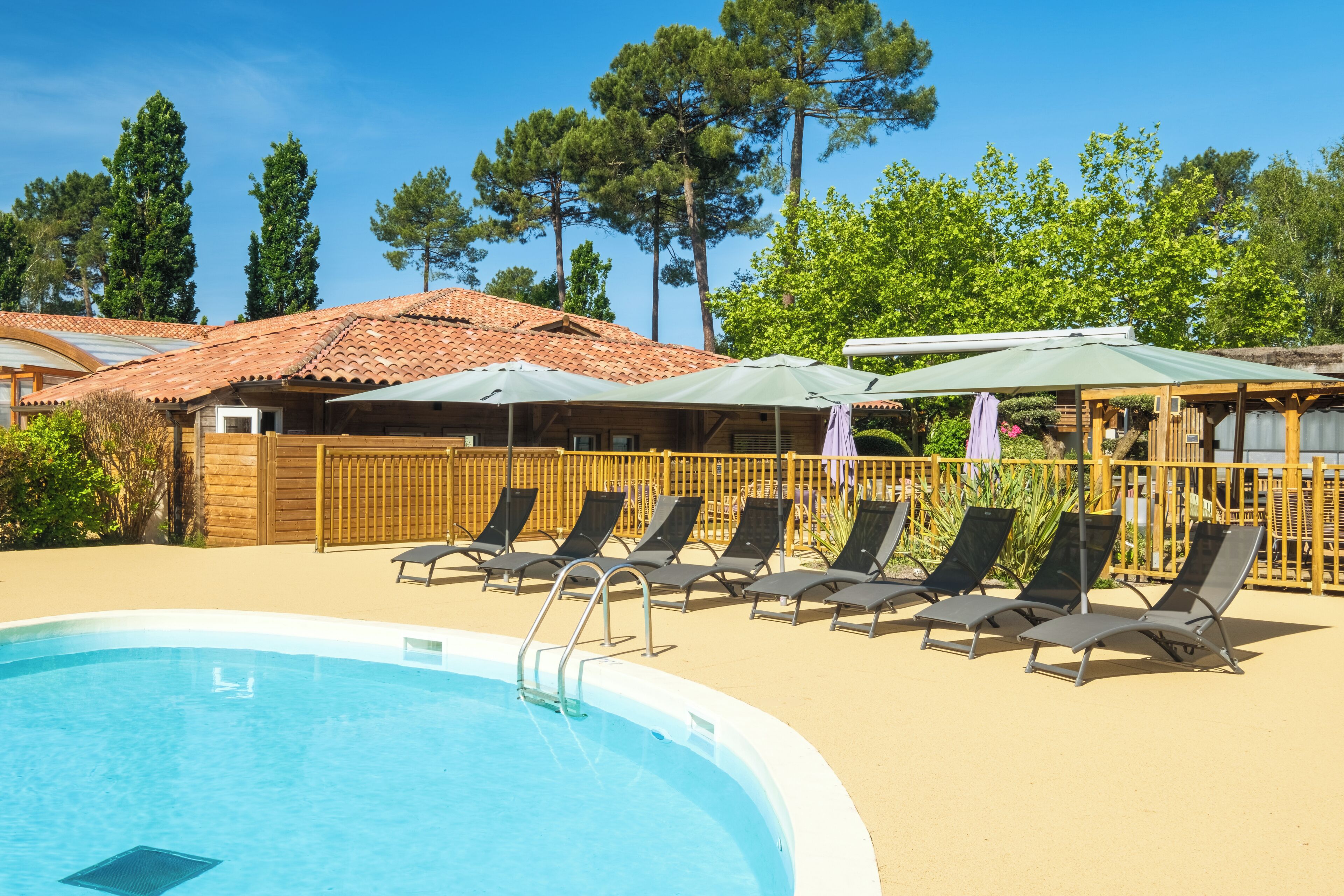 Una piscina con sillas de descanso y sombrillas, rodeada de vallas de madera y árboles.