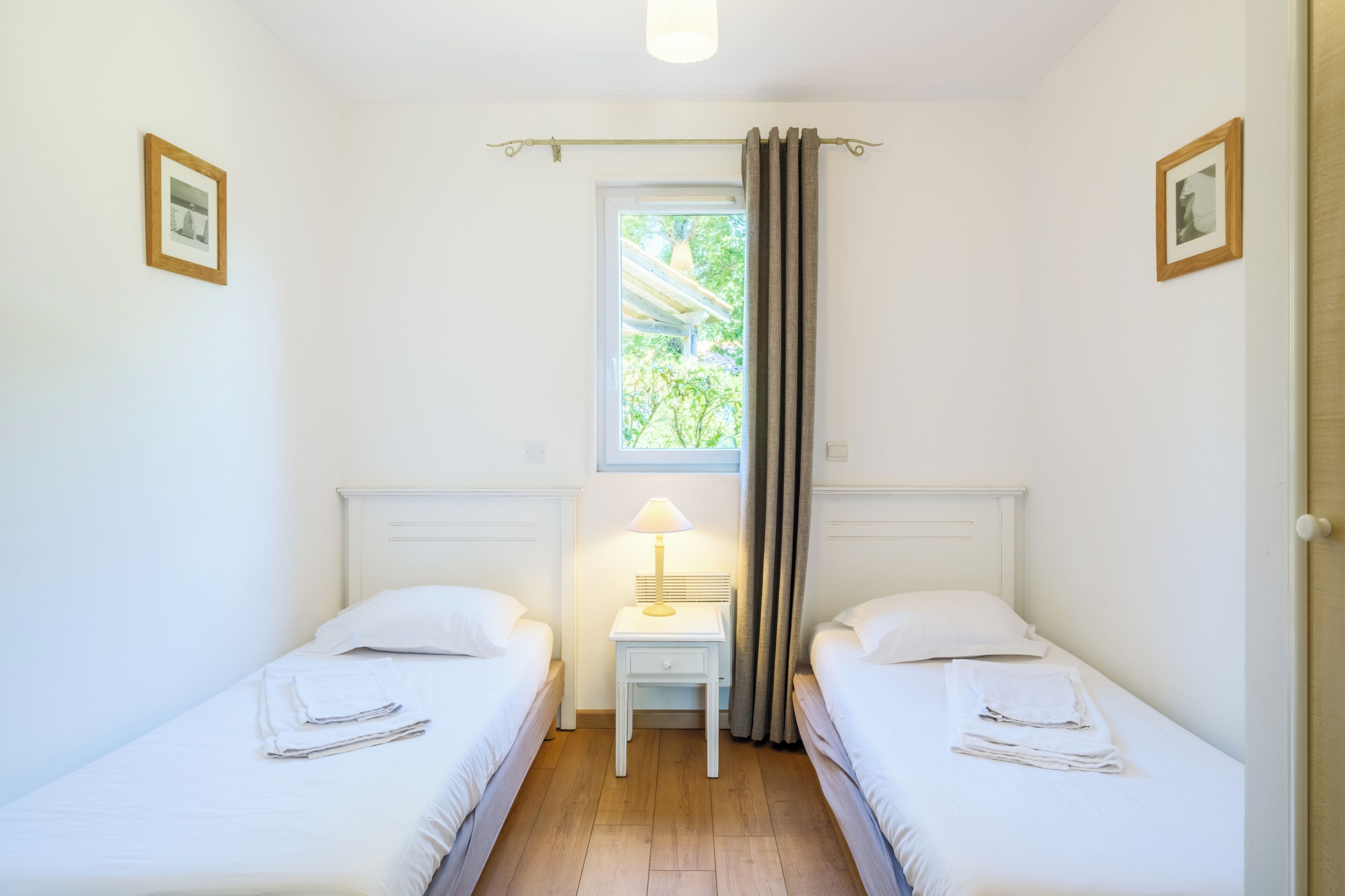 A twin room with two beds, a small table with a lamp, a window with curtains, and framed pictures on the walls.