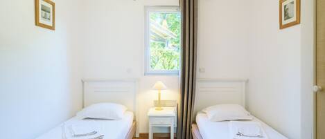 A twin room with two beds, a small table with a lamp, a window with curtains, and framed pictures on the walls.