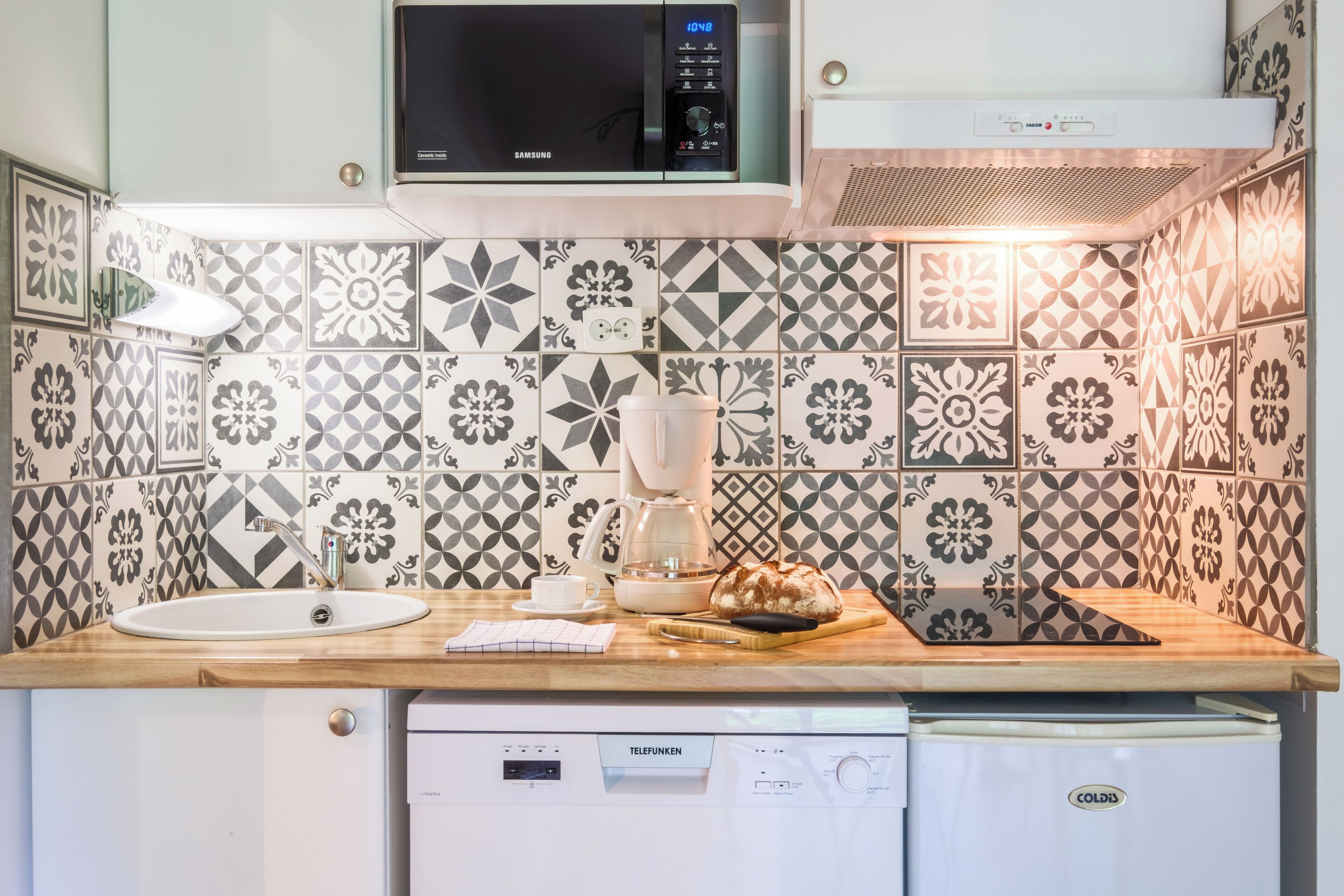 A kitchen with patterned tiles, a microwave, a coffee maker, a bread loaf, and a dishwasher.