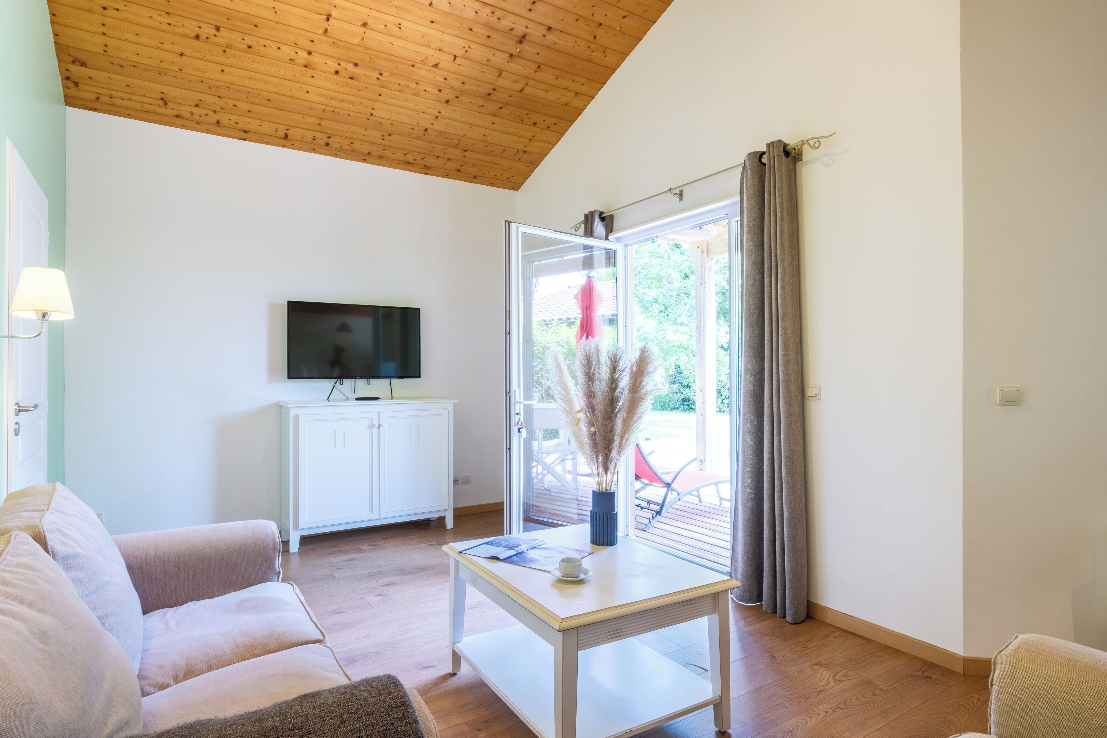 A living room with a television on a white cabinet, a coffee table with a book and a cup, a sofa, and a sliding glass door leading to an outdoor area.