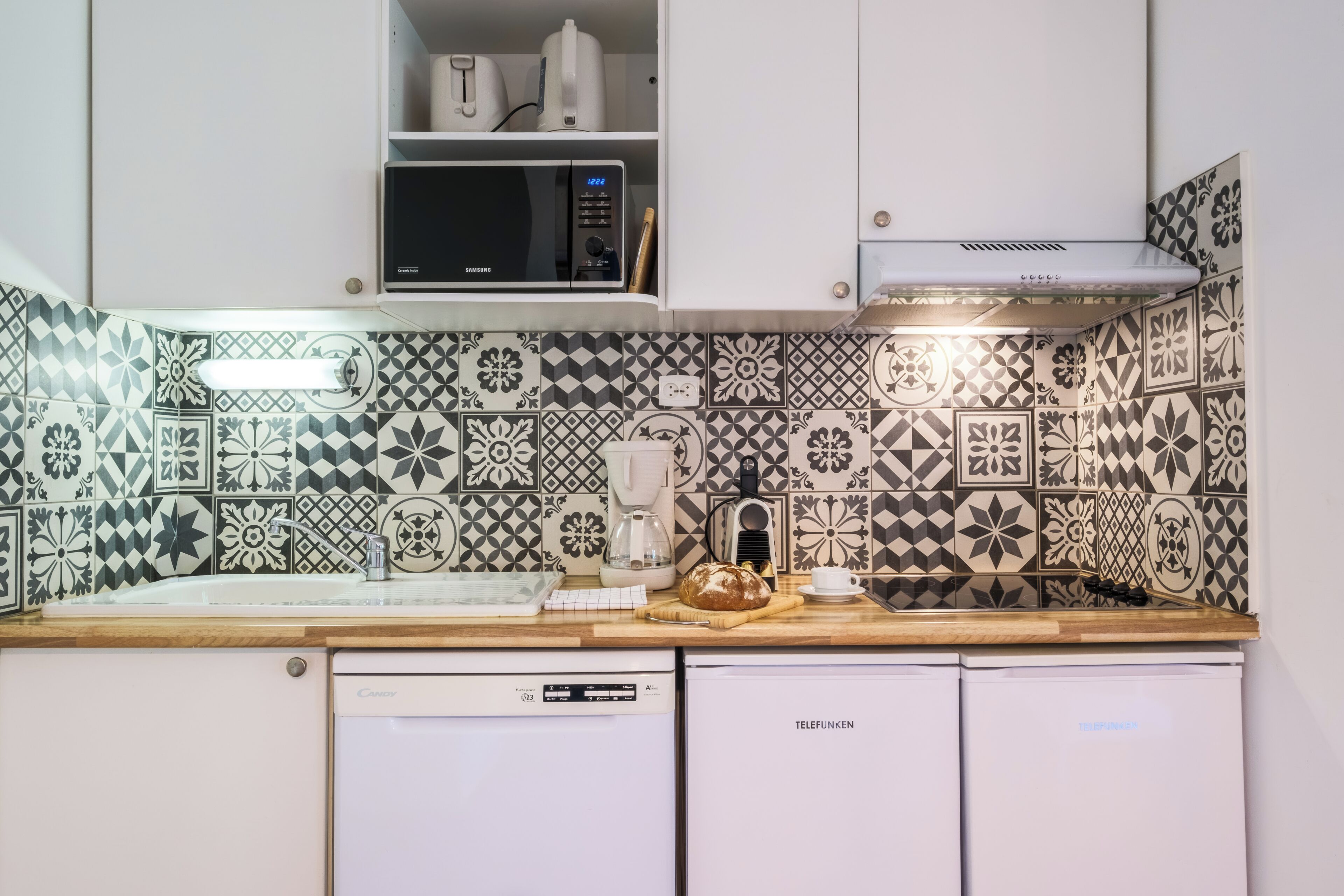 A kitchen with patterned tiles, a microwave, a coffee maker, a bread loaf, and a dishwasher.